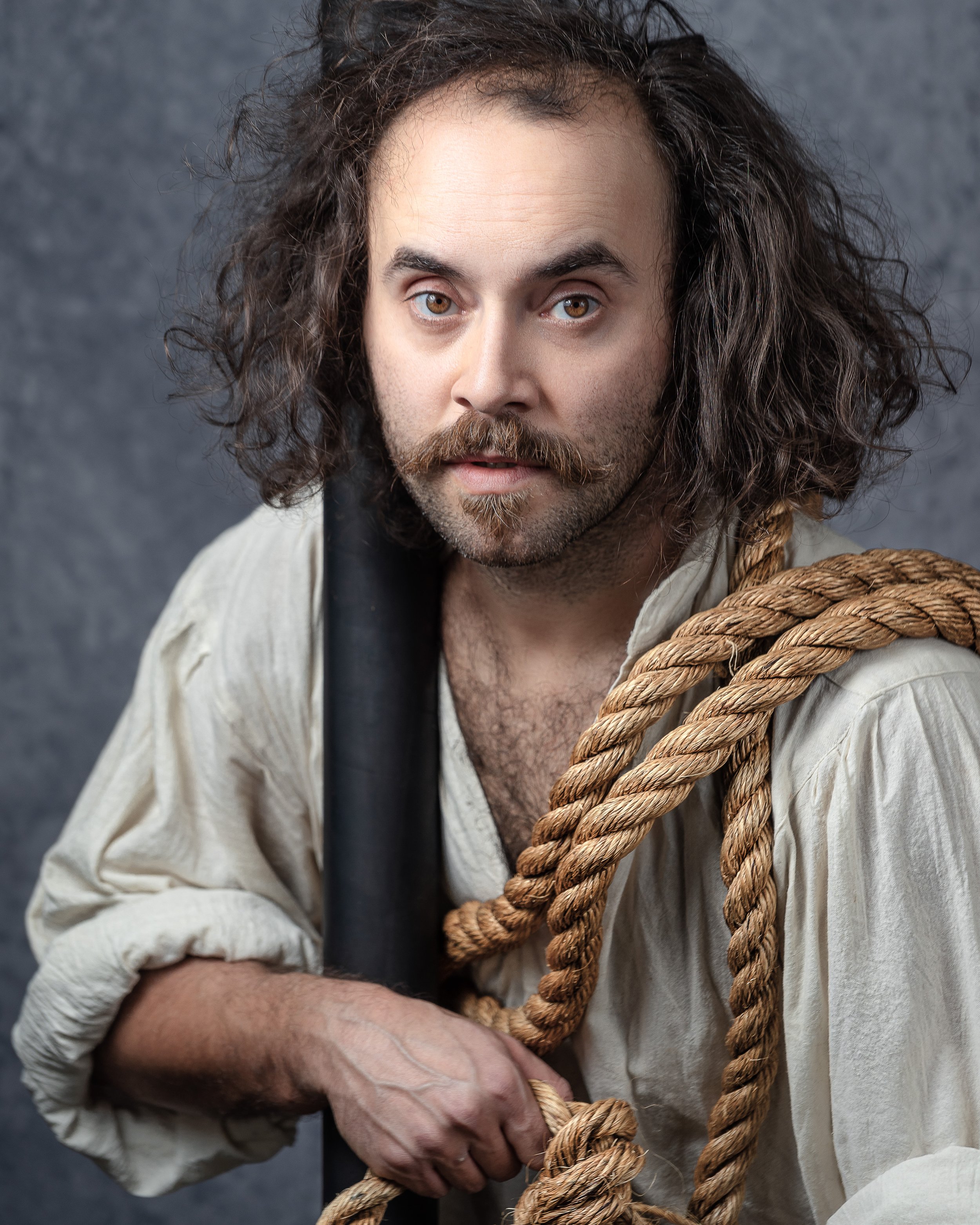 A man with long curly hair, a beard, and light brown eyes, wearing a beige shirt and a dark vest, is gripping a thick rope.