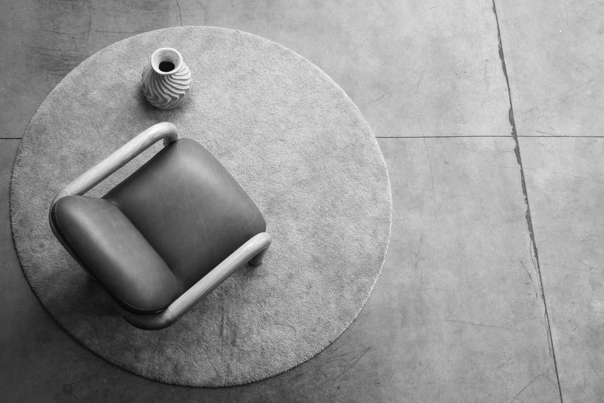 A top-down view of a leather armchair with light wooden armrests placed on a round textured rug. Next to the chair, on the rug, is a small decorative vase with a swirling pattern, holding a dark substance, possibly a flower vase, in a minimalist setting.