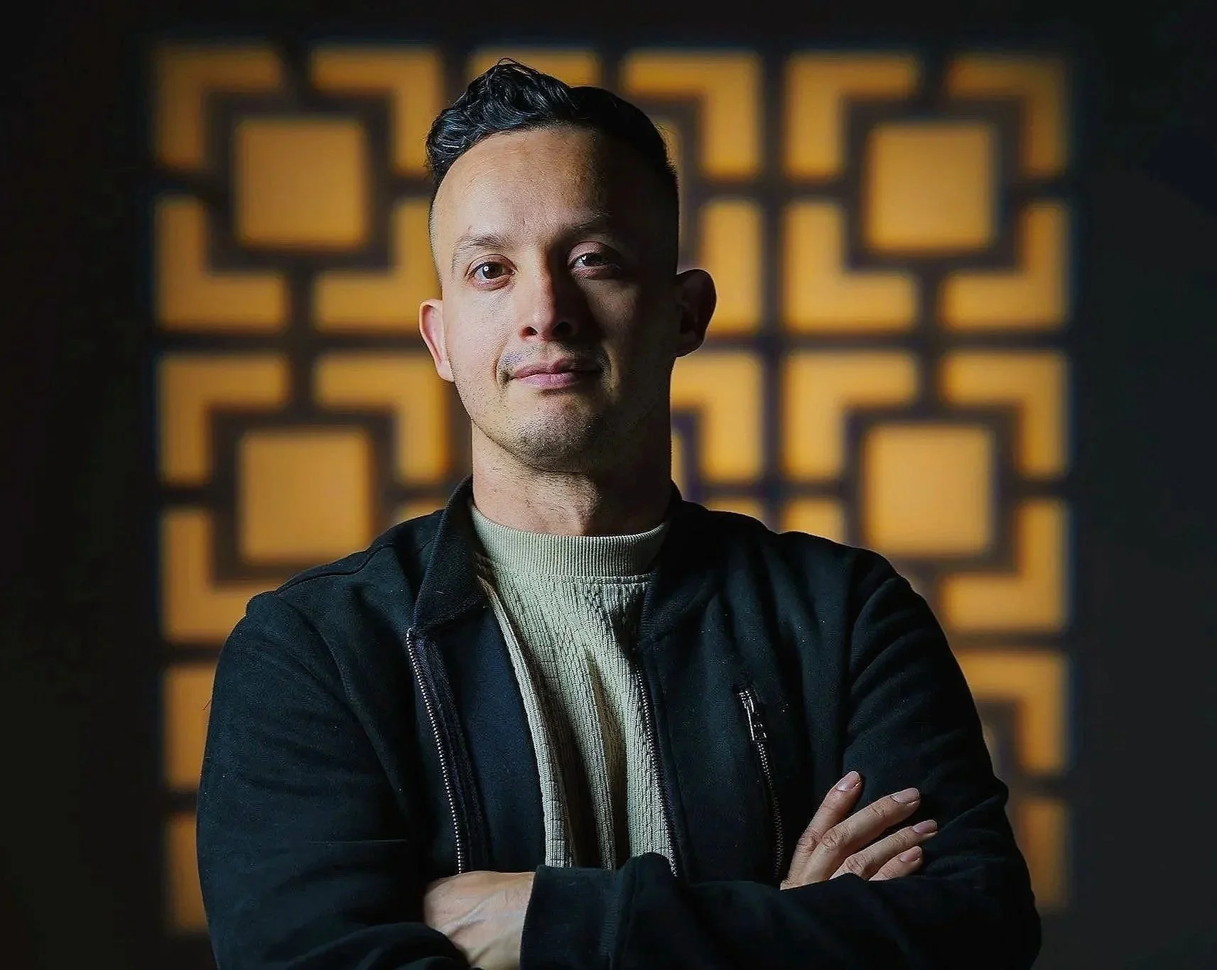 Portrait of a young man with short dark hair, wearing a black jacket over a beige sweater, standing with arms crossed in front of a geometric patterned background with warm lighting.