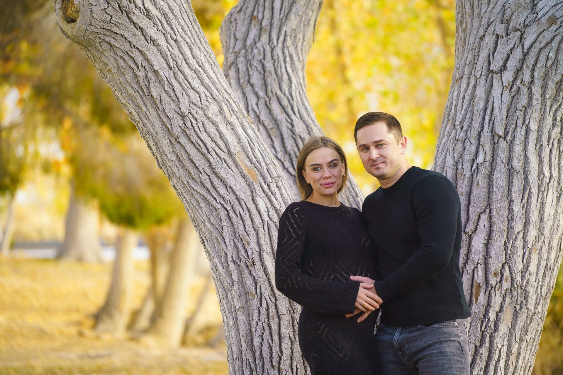 A couple stands close together outdoors in front of a tree with textured bark, surrounded by fall foliage in yellow and orange hues.