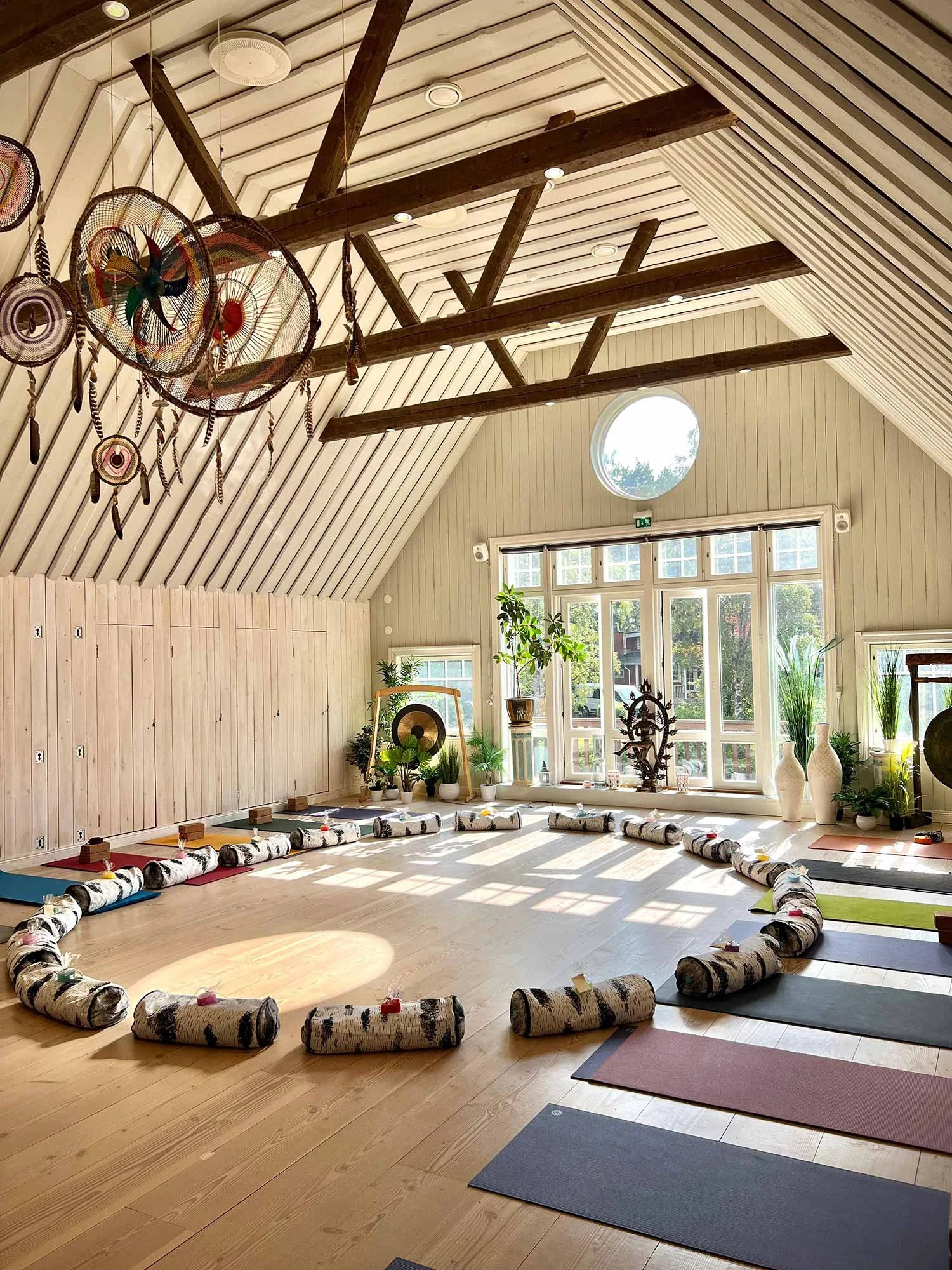 Villa Mandalan valoisa jooga- ja meditaatiotila auringonvalossa, lattialla on joogamattoja ja suuren ikkunan edessä useita viherkasveja.