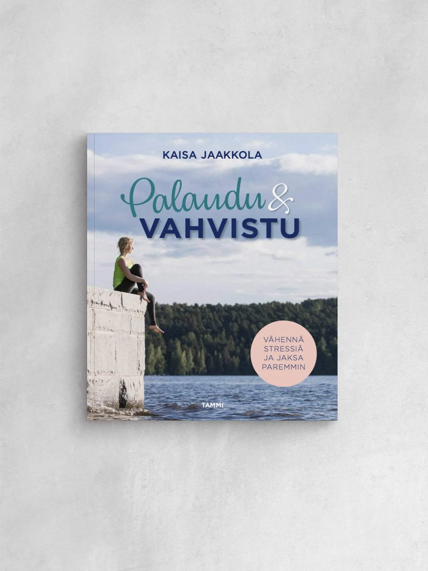 Kirjan kansi, jossa nainen istuu betonikantaterassilla joen rannalla, taustalla puita ja pilviä, otsikko 'Palaudu ja Vahvista' ja teksti 'Vähennä stressiä ja jaksa paremmin'.