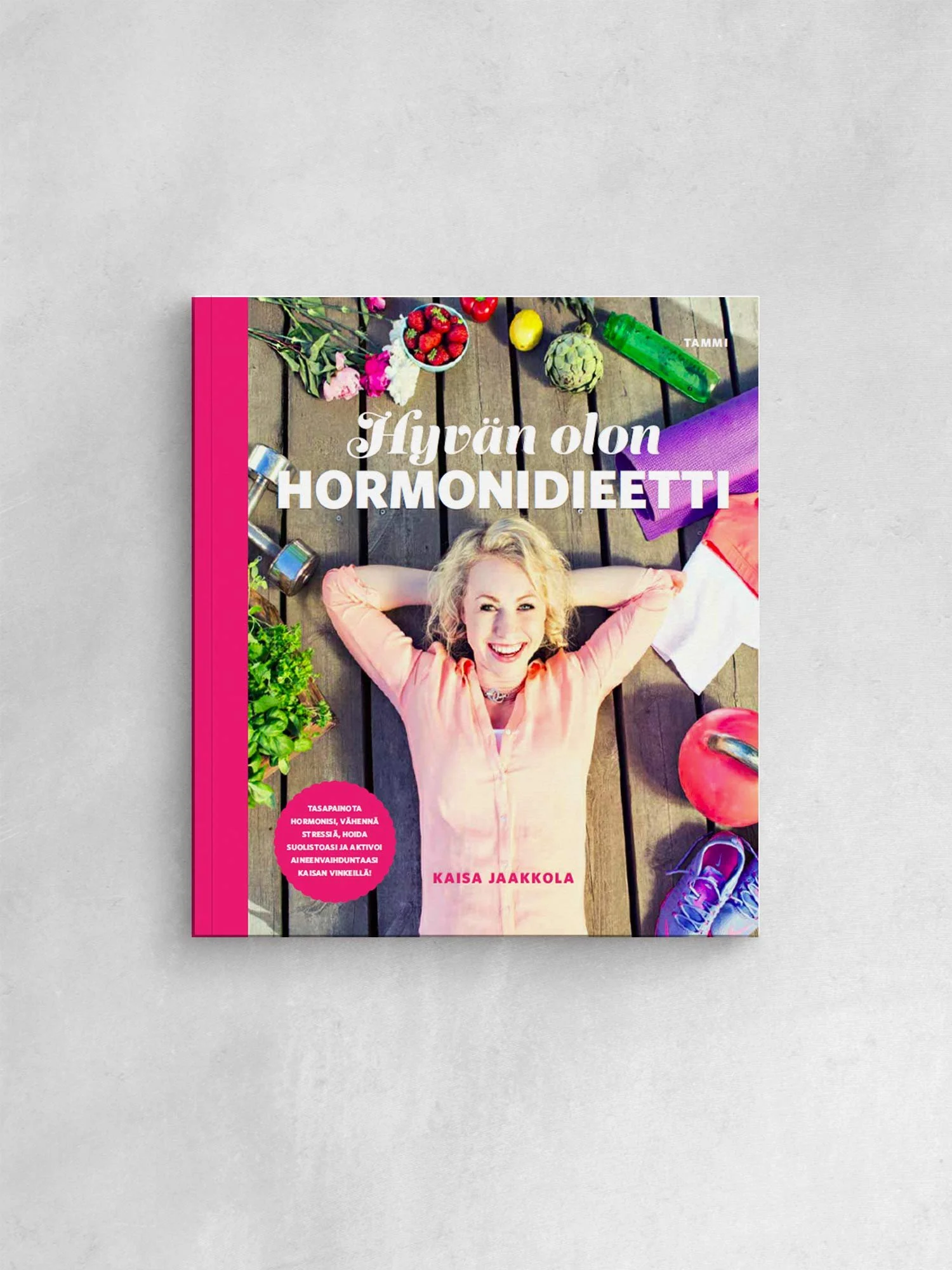 Värikäs kirja, jonka nimi on 'Hyvän olon Hormonideetti' ja kannessa on nainen, joka makaa selällään puulattialla, hymyilee ja tuo kädet pään taakse, ympärillä on kasveja, hedelmiä ja urheiluvälineitä.