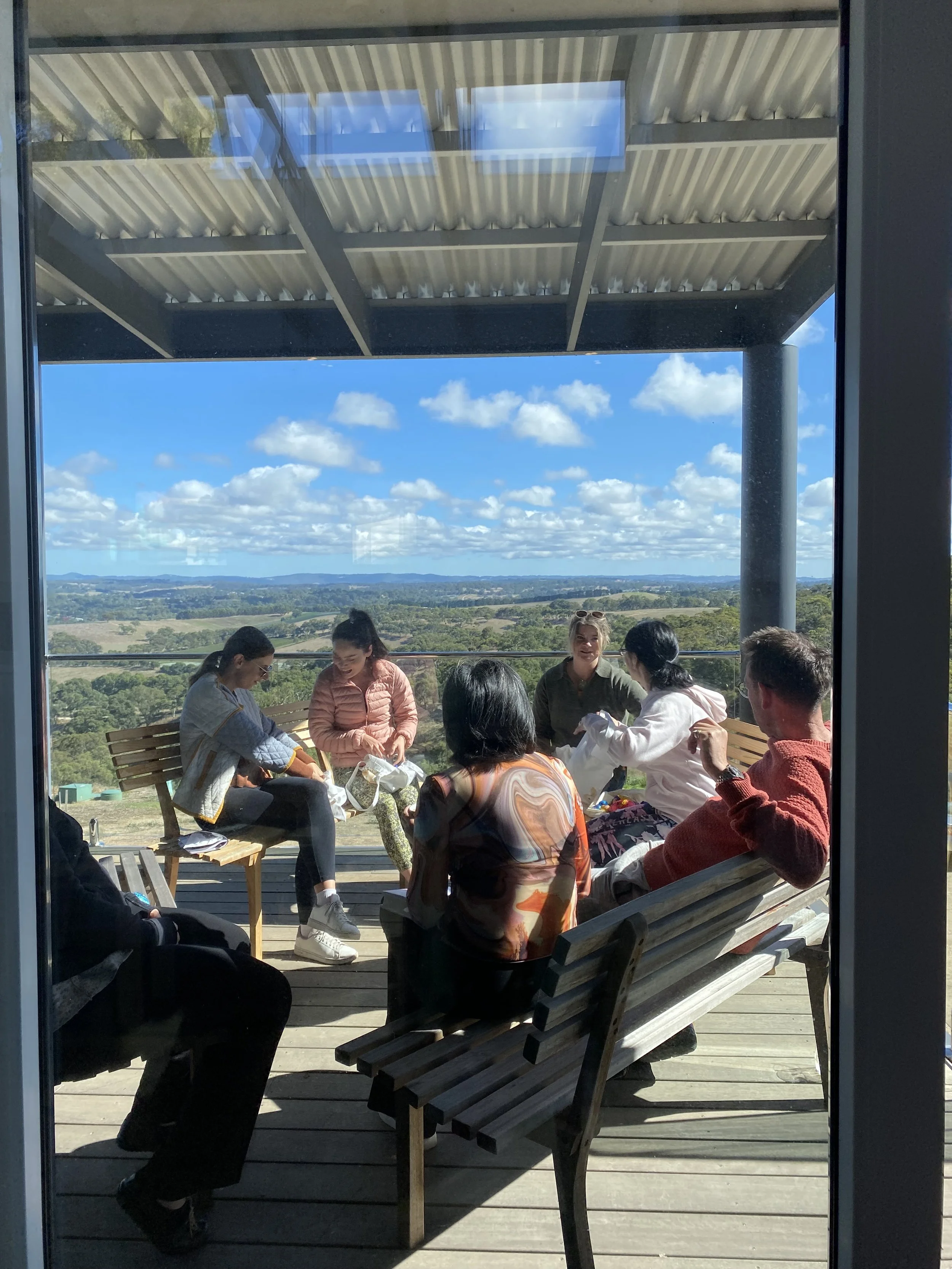 A group of seven people sitting outdoors on a wooden deck, engaged in a discussion. They are seated on wooden benches and chairs with cushions. The setting offers scenic greenery and a view of the landscape in the background, with some people having notebooks or laptops.