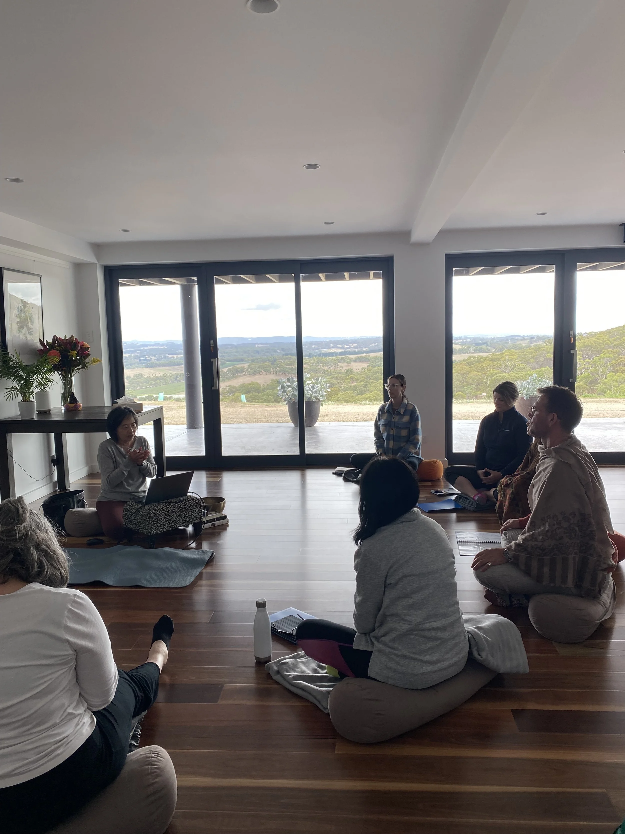 A group of people participating in a meditation class in a well-lit room with large windows, sitting on yoga mats in a cross-legged position.