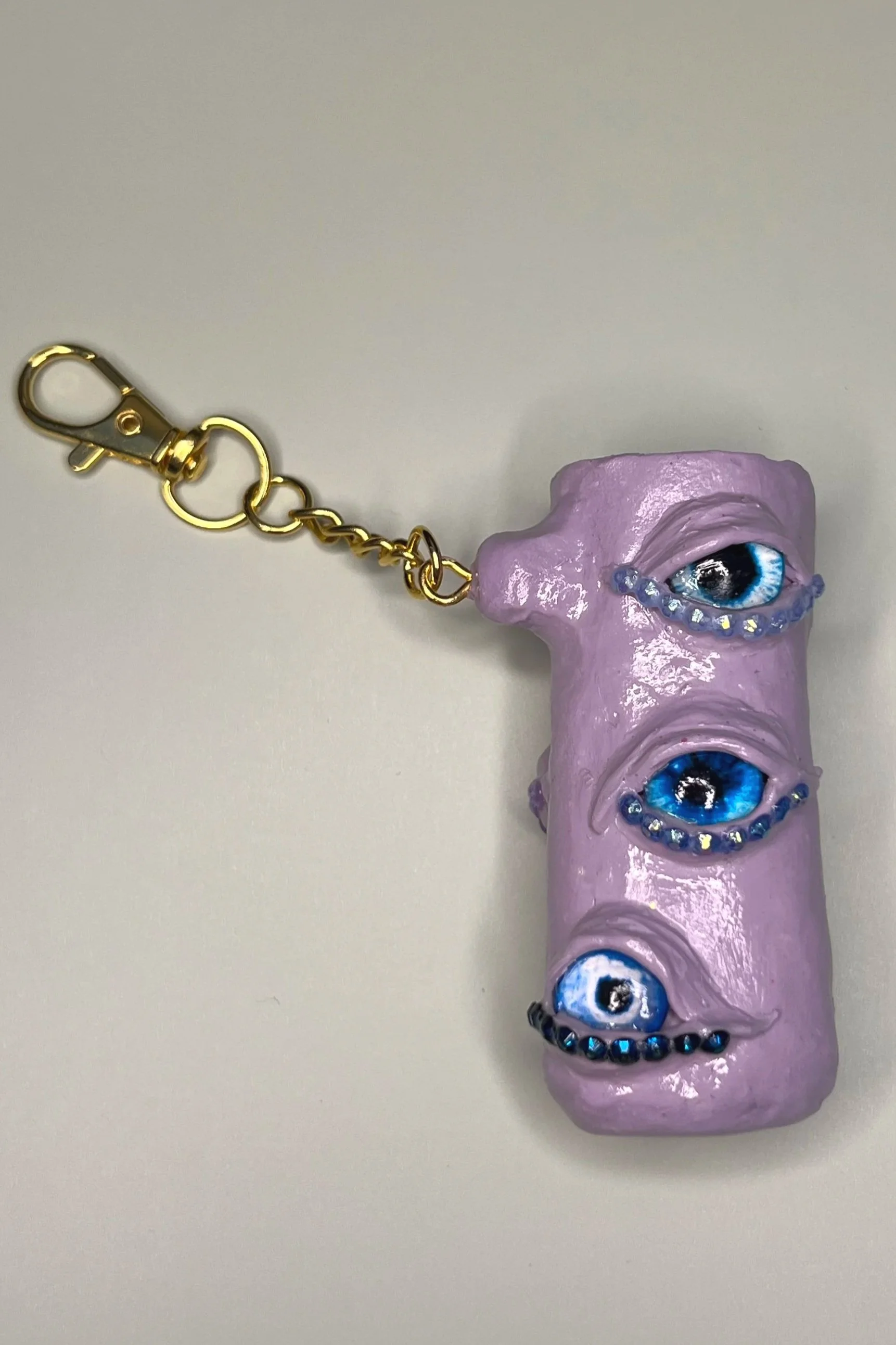 A purple lighter case keychain with three realistic blue eyes and rhinestones, attached to a gold chain with a clip.