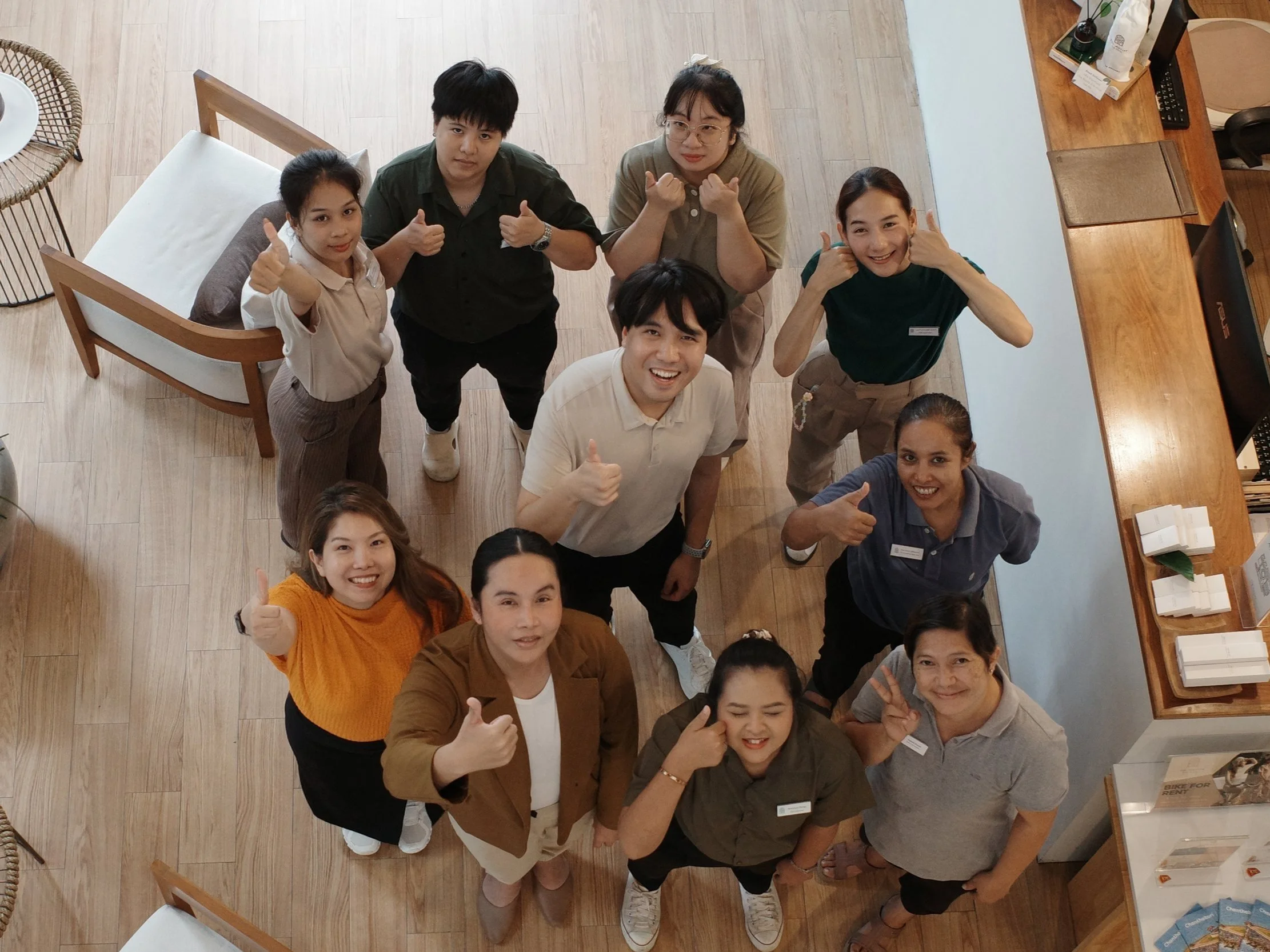 Group of people smiling and giving thumbs up, looking up at the camera in a room with wooden floors and furniture.