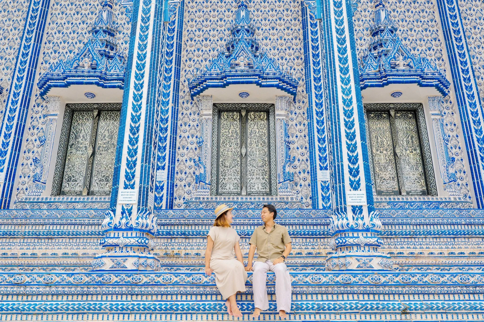 A man and woman sitting on steps in front of Wat Pak Nam Kam Noo, Chanthaburi. A blue and white ornate building with intricate patterns and windows.
