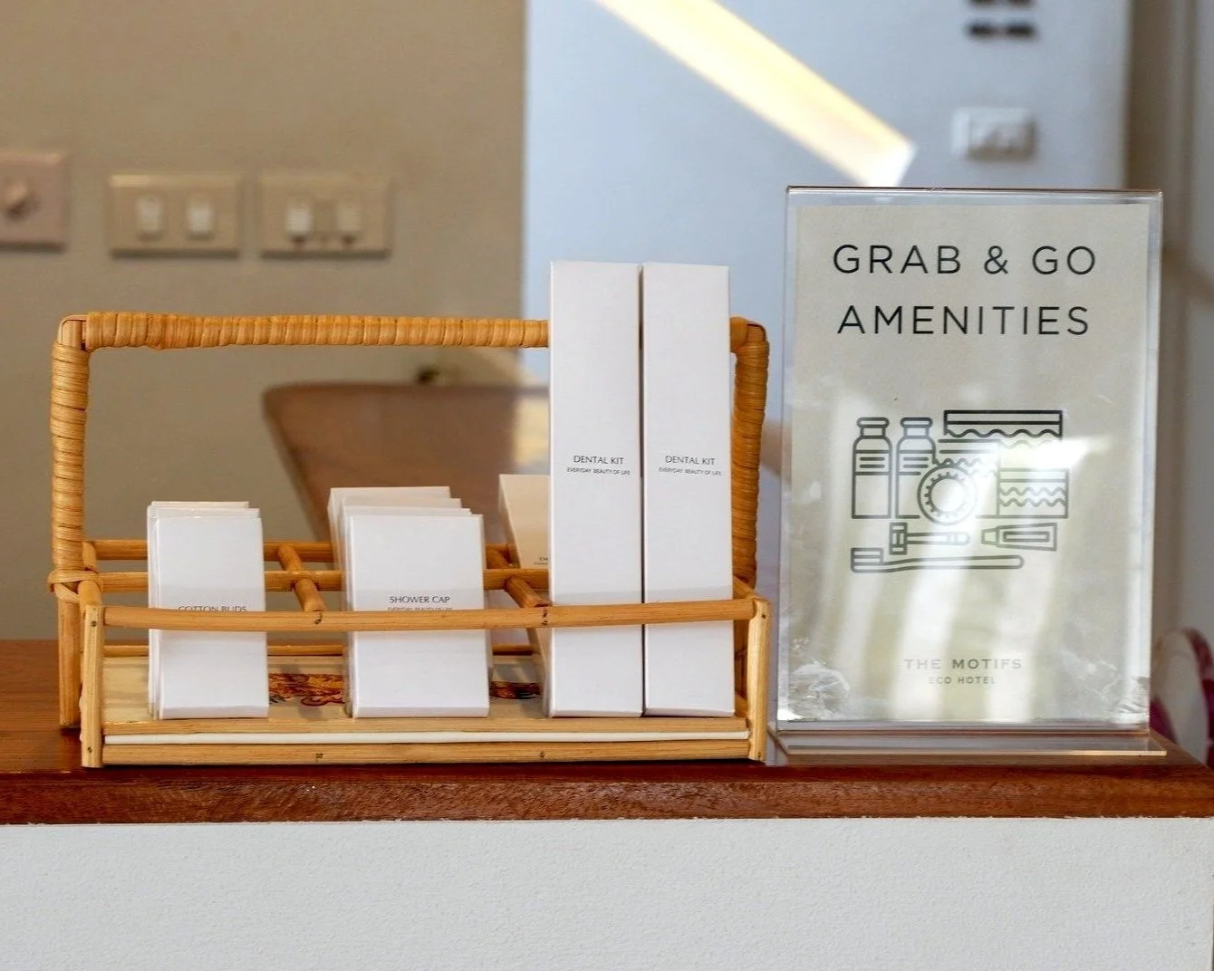 Travel amenities including dental kits, shower caps, and cotton buds in a wicker basket with a sign reading "Grab & Go Amenities" at a hotel reception desk.