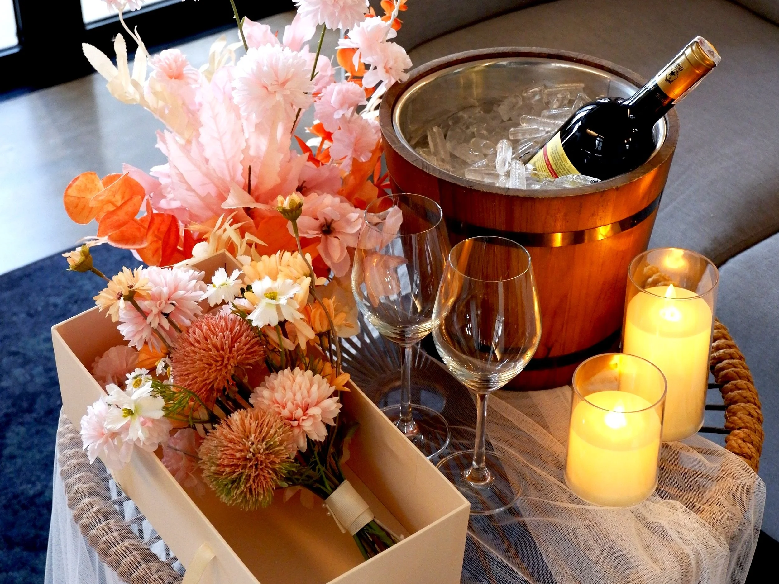 A cozy drink setup with a bottle of wine in an ice bucket, three wine glasses, lit candles, and a bouquet of pink and white flowers on a table covered with netted fabric. Perfect for honeymoon set up