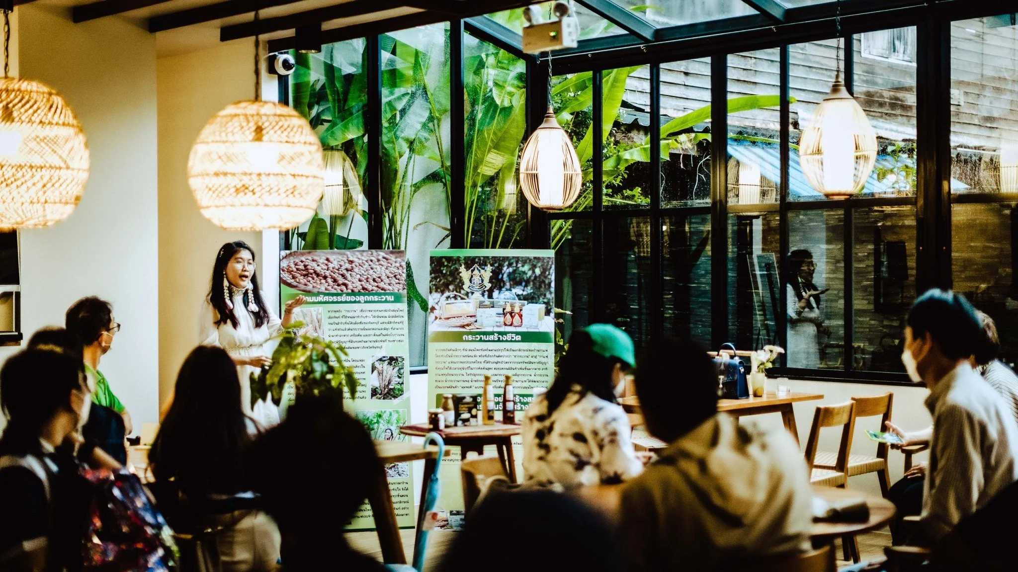 A group of people gathers indoors, listening to a woman giving a presentation next to display boards, with large windows showing greenery outside, and hanging pendant lights overhead.
