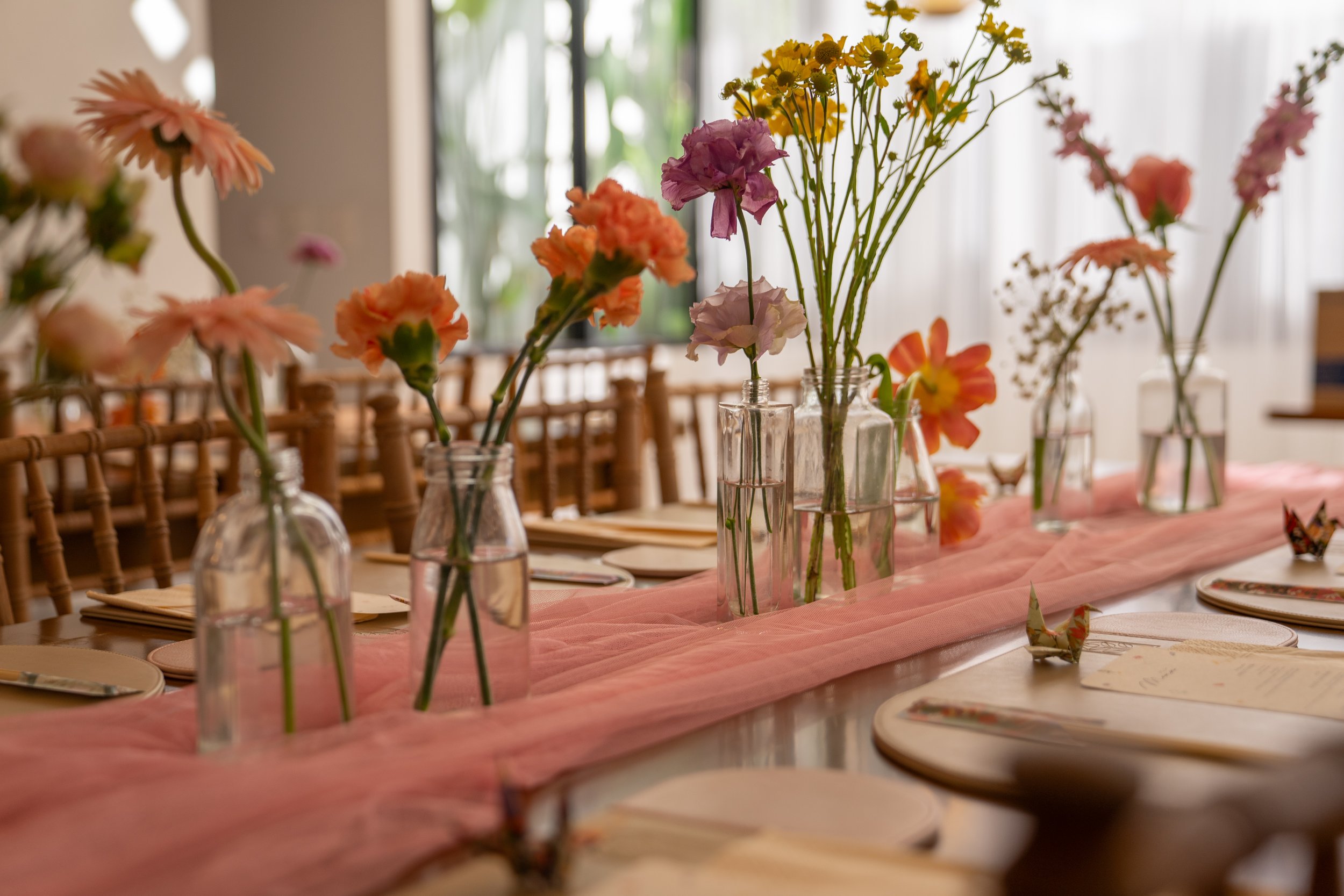 A table decorated with pink and peach flowers in various glass vases, with place settings and origami paper cranes, on a pink table runner in a well-lit indoor space.
