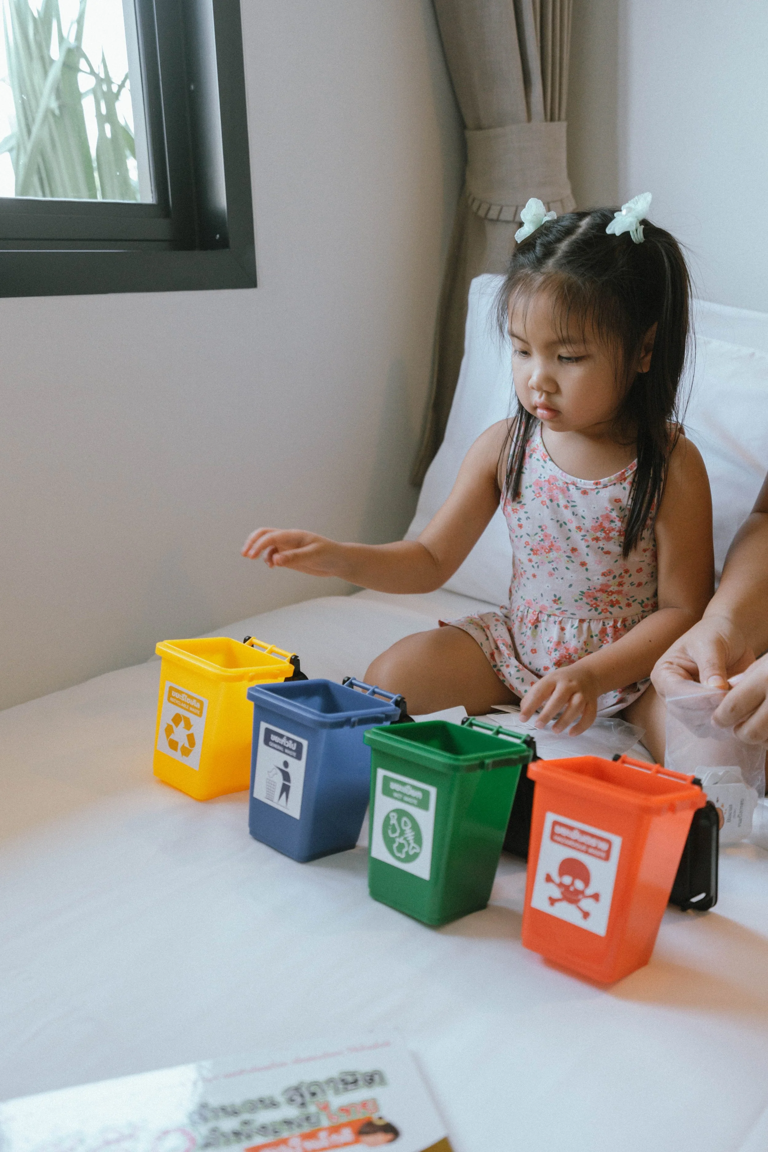 Young girl sitting on bed with four colored miniature recycling bins labeled for different waste types.