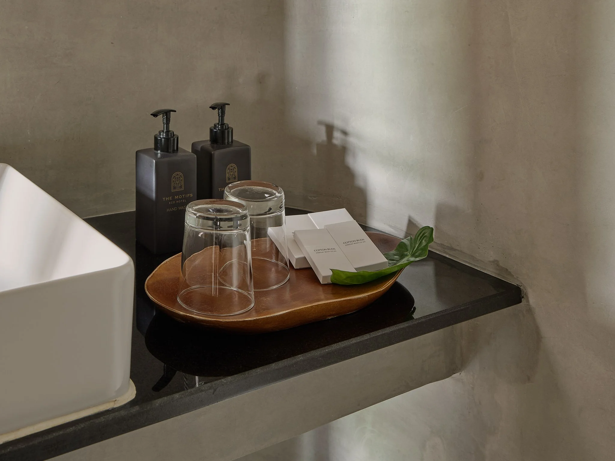A wooden tray holds two upside-down glass cups, three small white boxes labeled 'Cotton Buds', and a green leaf, placed on a black countertop with bottles of hand wash and a white sink nearby.