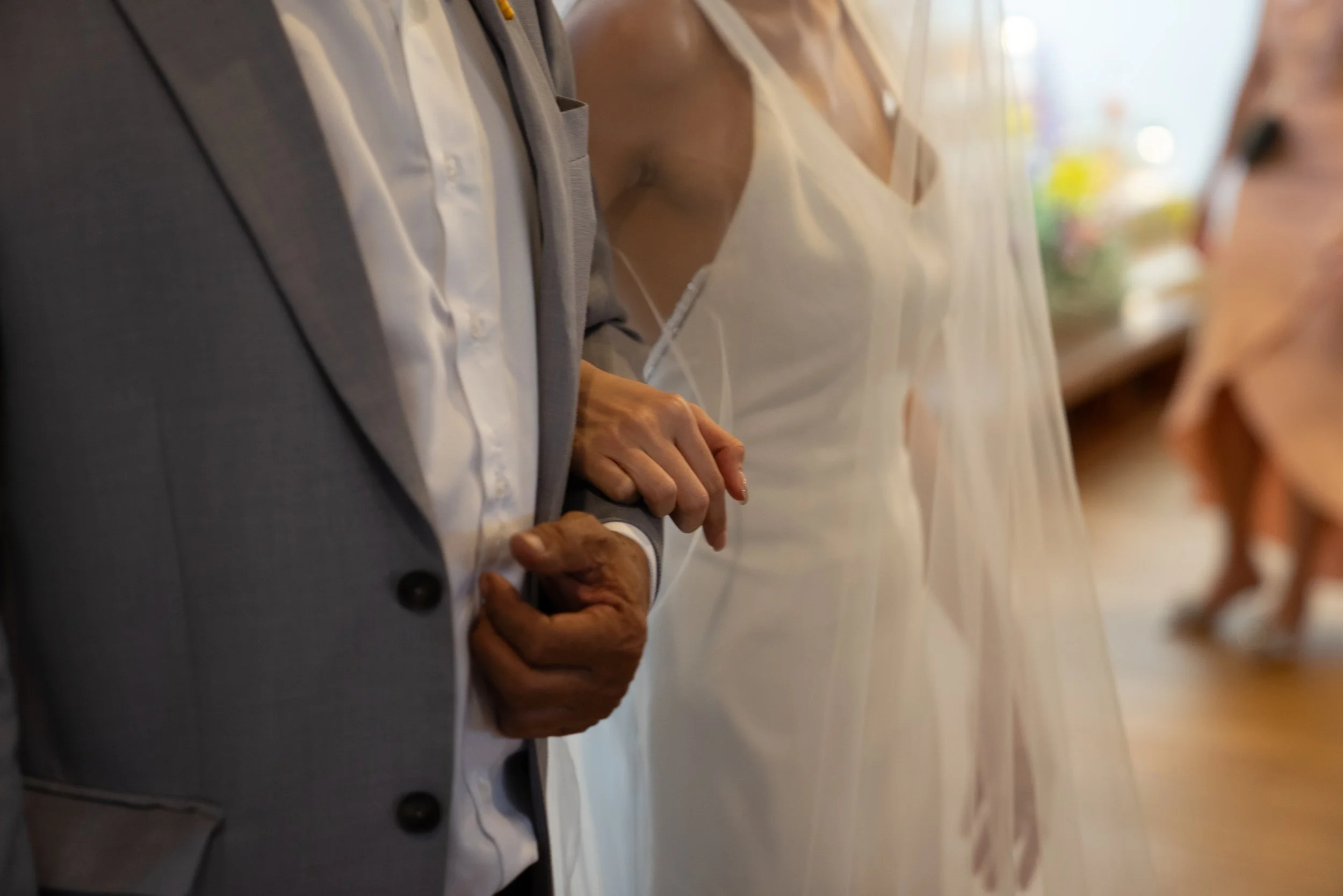 Close-up of a couple holding hands during a wedding ceremony, with the man in a gray suit and white shirt and the woman in a white wedding dress and veil.