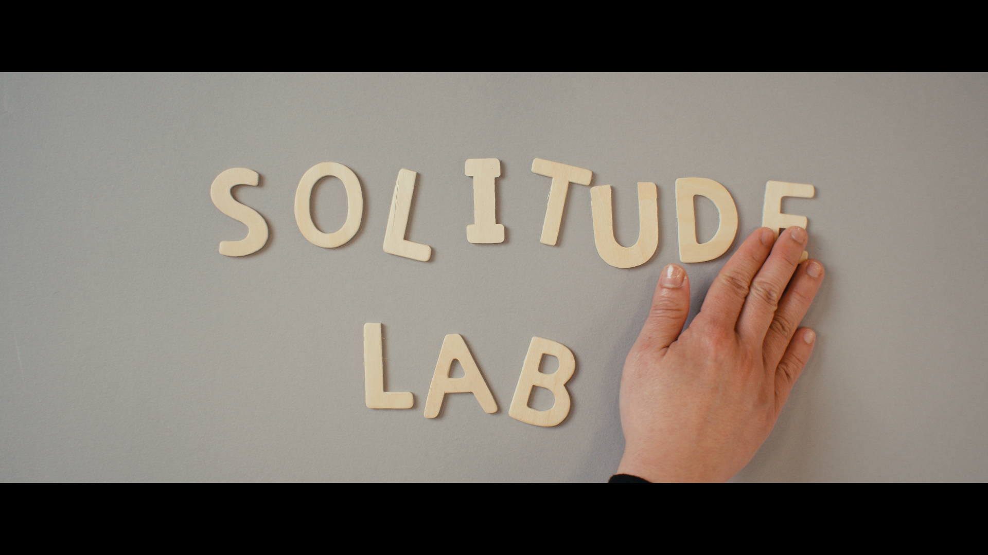 Hand arranging beige letter tiles on light gray background, spelling 'SOLITUDE LAB'.