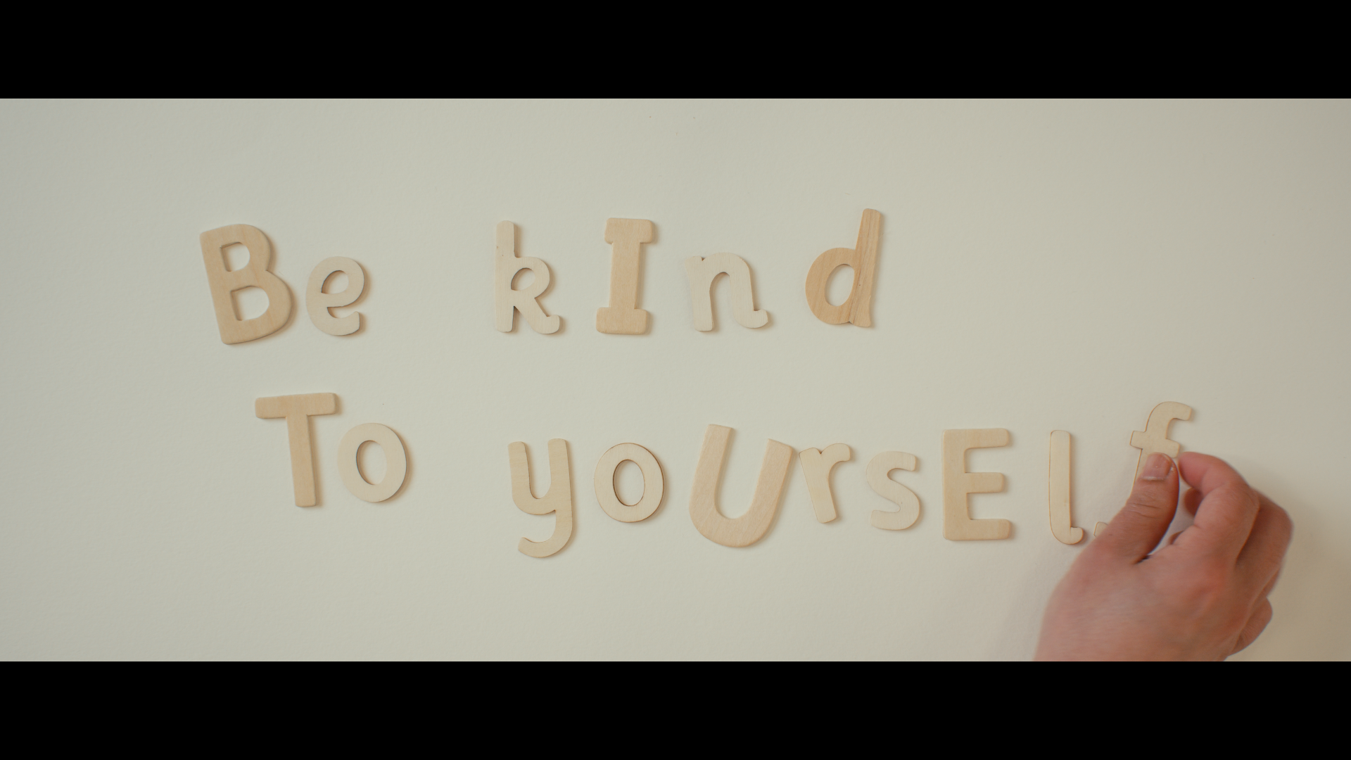Wooden letter tiles on a white background spell out 'Be kind to yourself,' with a hand in the lower right corner placing the letter 'f'.