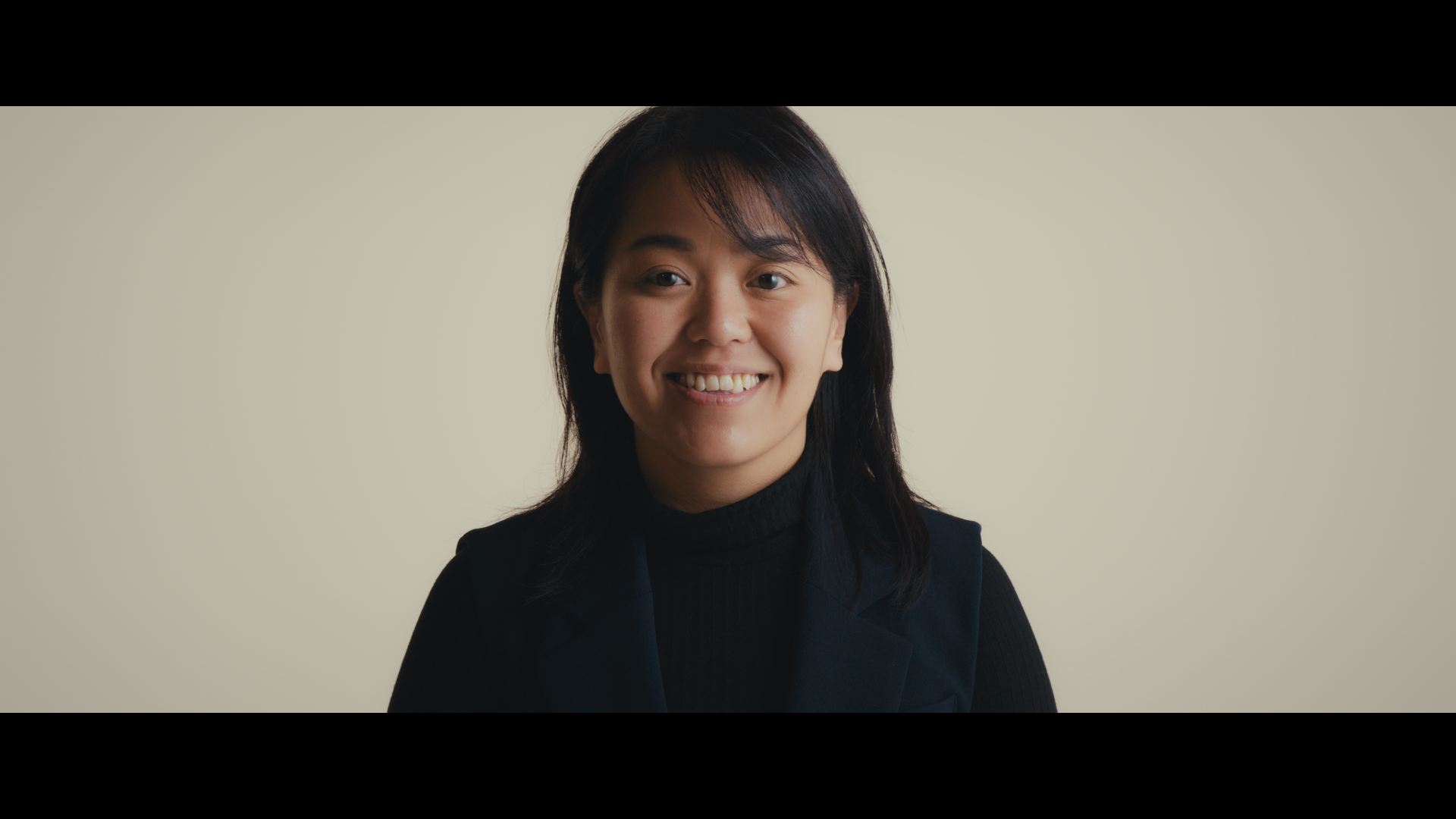 A woman with shoulder-length dark hair smiling against a plain light-colored background, wearing a black top and vest.