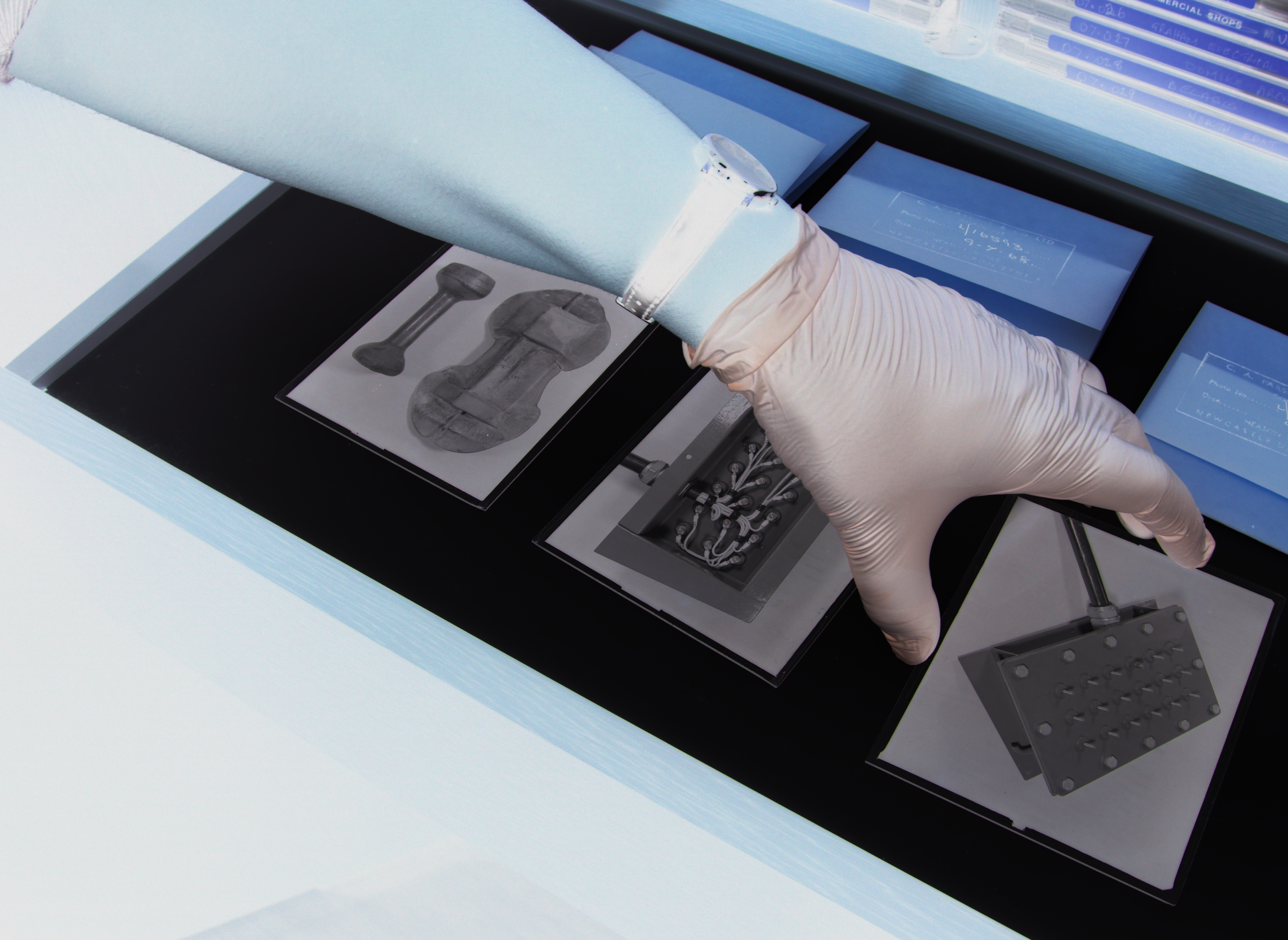 A gloved hand pointing to a petri dish with a metallic structure on a black surface, surrounded by scientific equipment and blue folders.