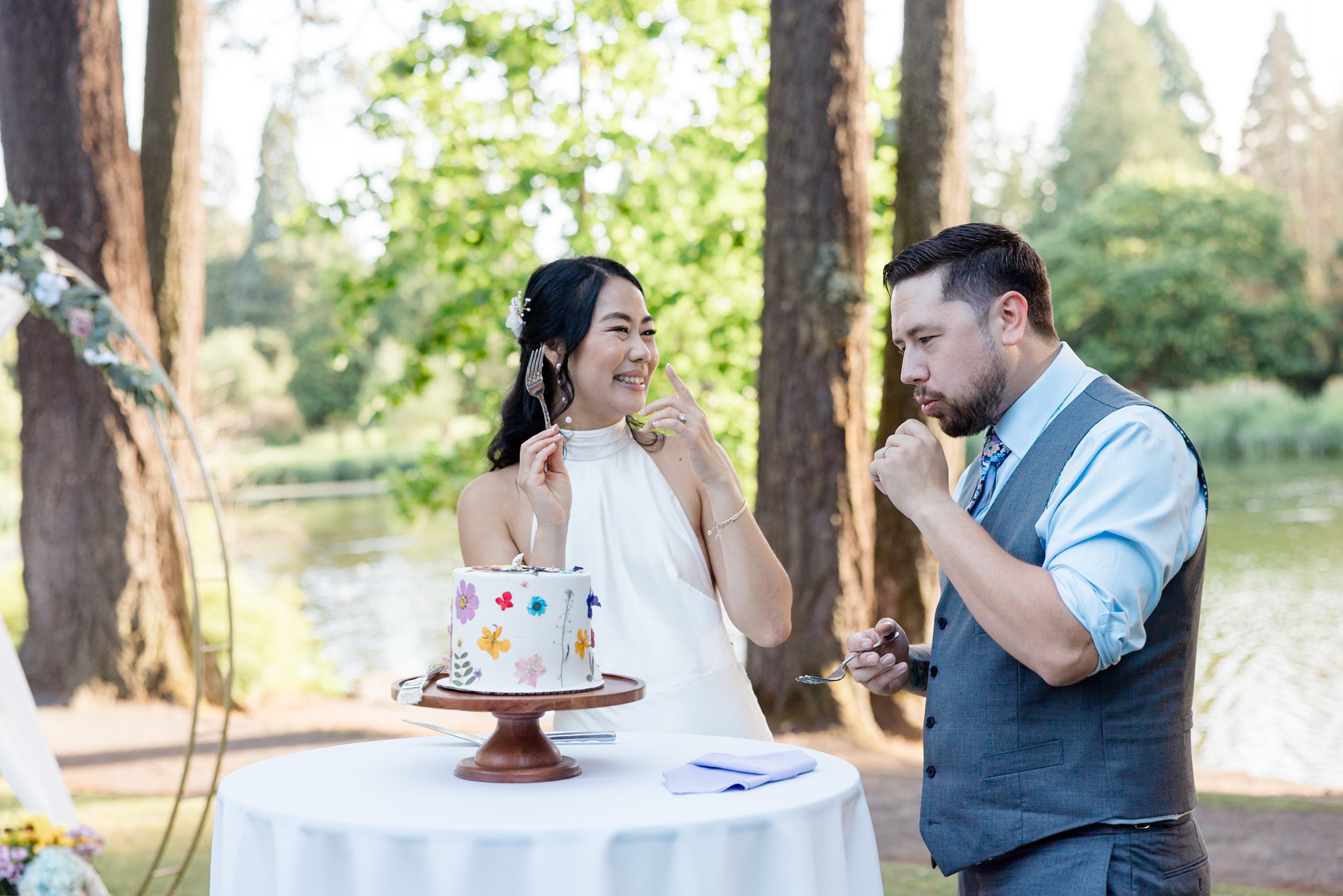 A couple celebrating with a decorated cake at an outdoor event near a lake, with trees in the background. The woman is in a white dress and the man is in a gray vest and blue shirt. They are smiling and holding forks.