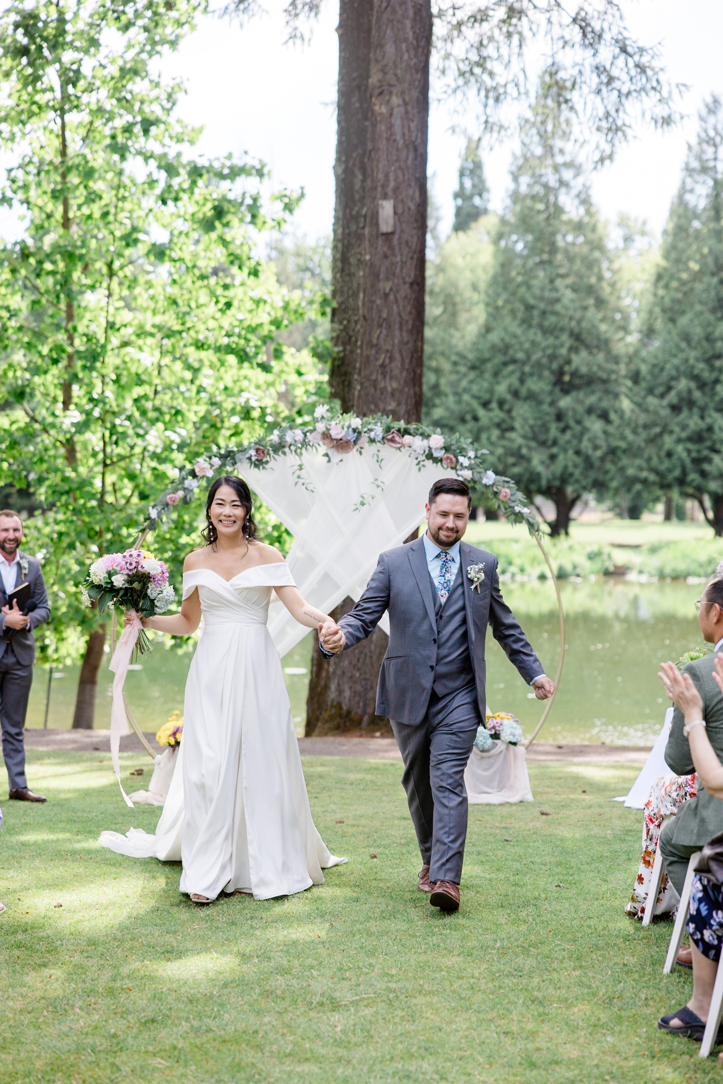 Bride and groom walking down a grassy aisle in an outdoor wedding ceremony. The bride is in a white gown holding a bouquet, and the groom is in a suit. An arch decorated with flowers and fabric is in the background, with trees and a lake visible.