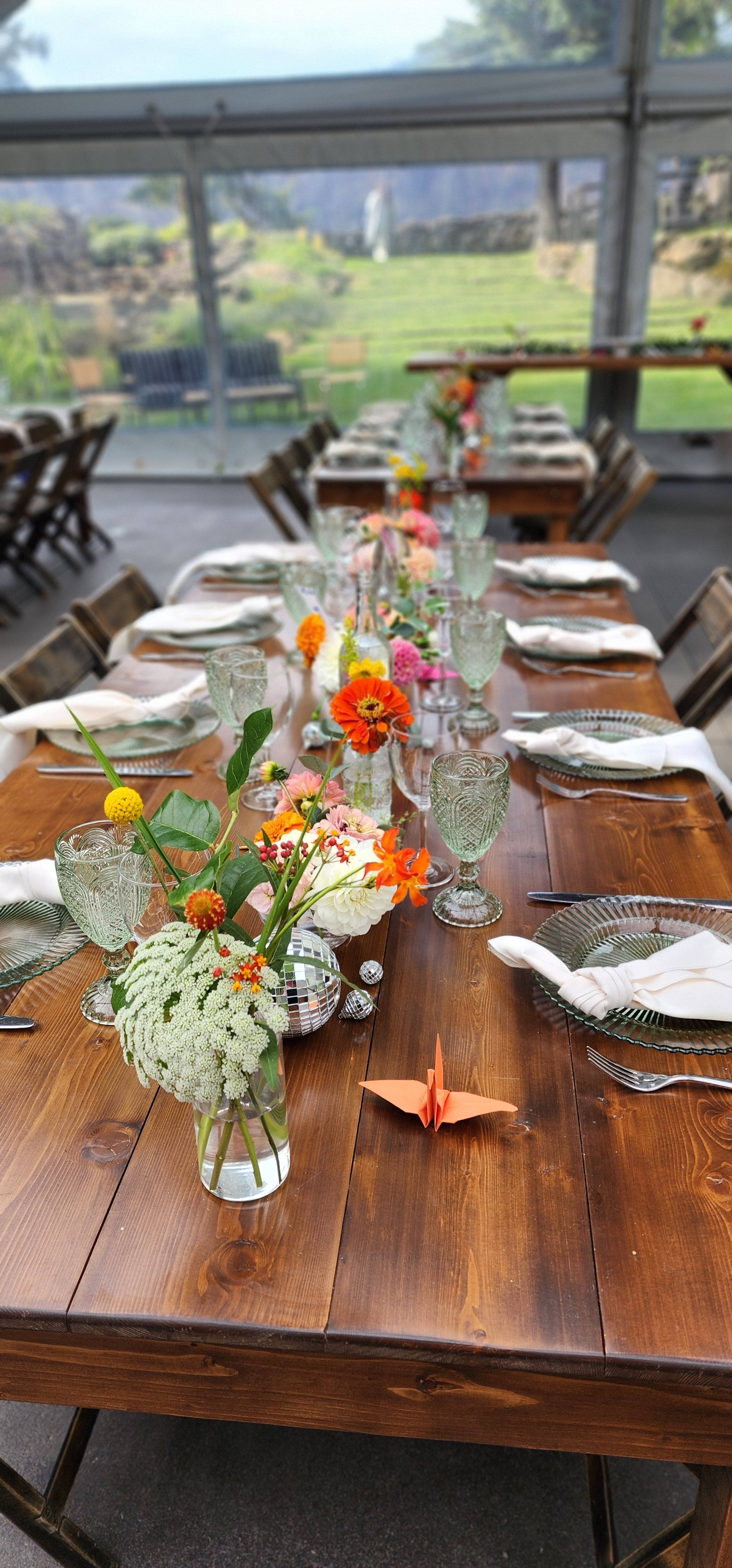 Elegant dining table setup with wooden table, glass plates, ornate glasses, white napkins, and floral arrangements in vases, featuring bright orange and white flowers. An orange origami crane decorates the table.