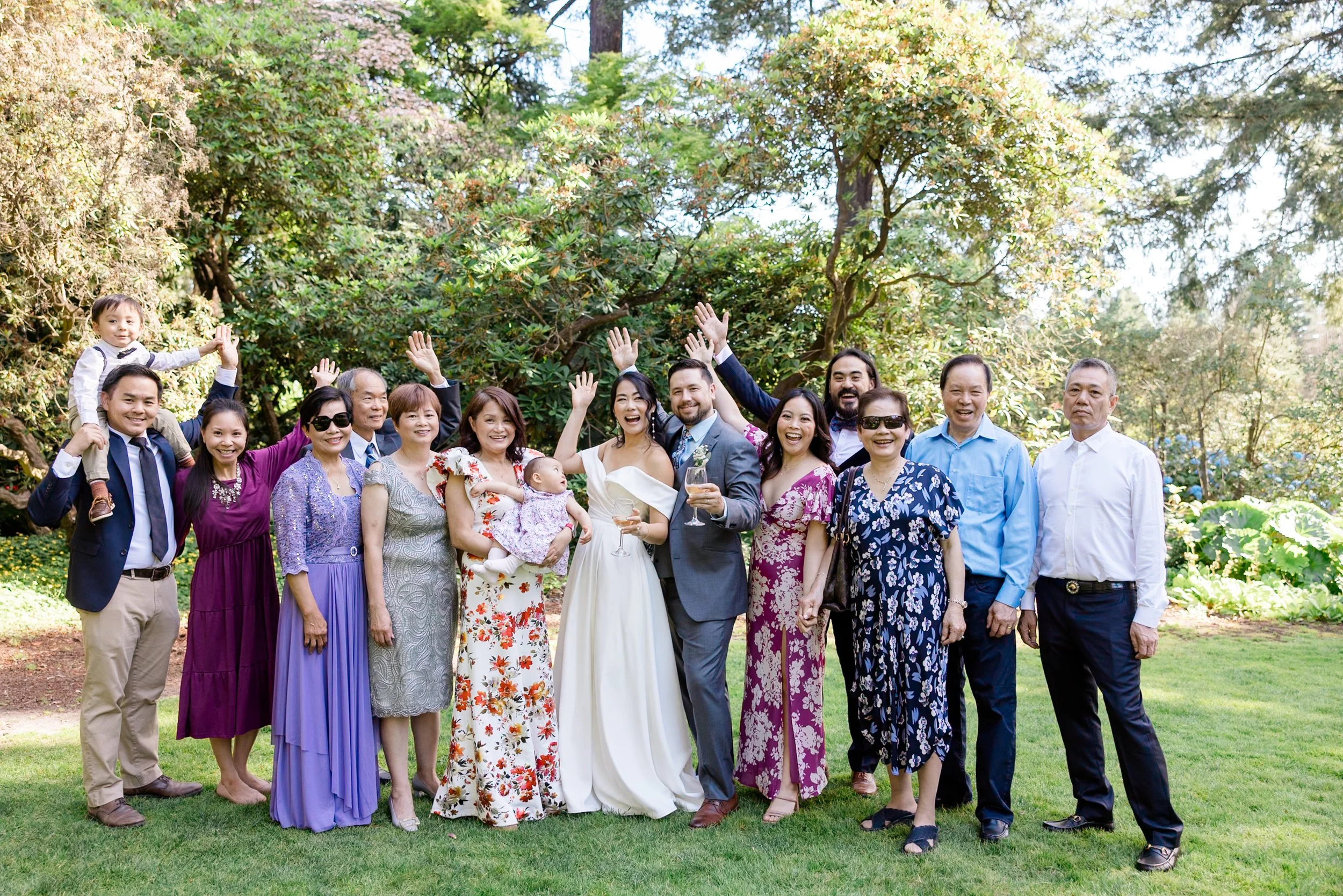 Group of people smiling and raising hands in a garden setting, including a bride and groom, various adults, a child, and a baby.