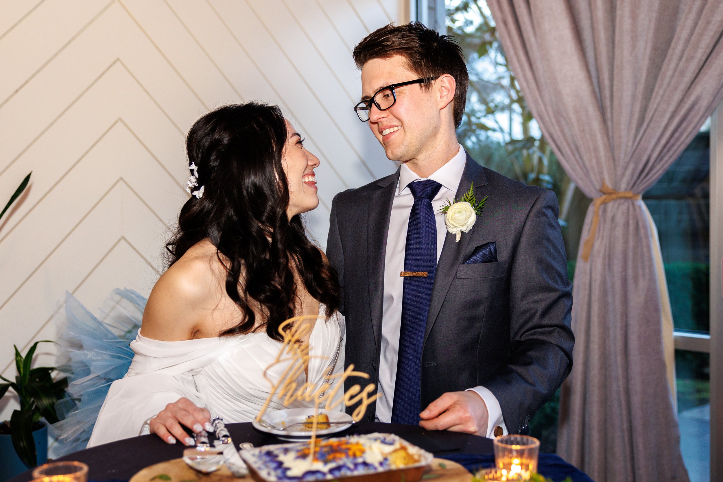 A bride and groom smiling at each other during their wedding reception, standing by a table with a decorative cake topper reading "The Huxleys." The groom is wearing a suit with a boutonniere, while the bride is in an off-shoulder white dress.