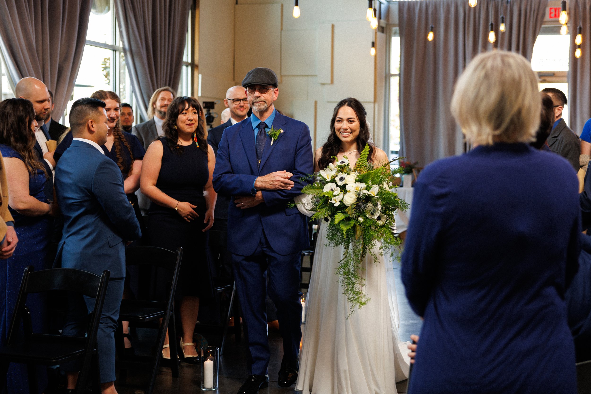 A bride in a white dress holding a bouquet is walking down the aisle with a man in a blue suit at a wedding ceremony. Guests are seated and standing around, watching the procession. The venue has dim lighting and curtains hanging on the windows.