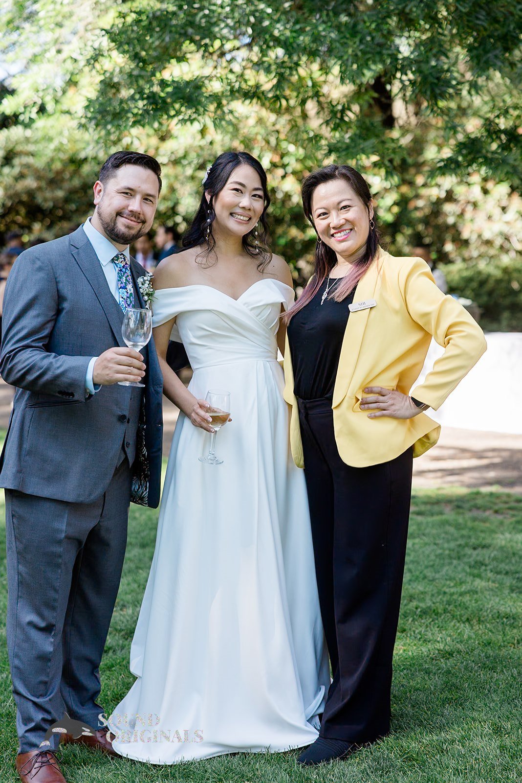 Three people at a wedding, with the bride in a white gown, standing on grass with trees in the background, holding wine glasses.