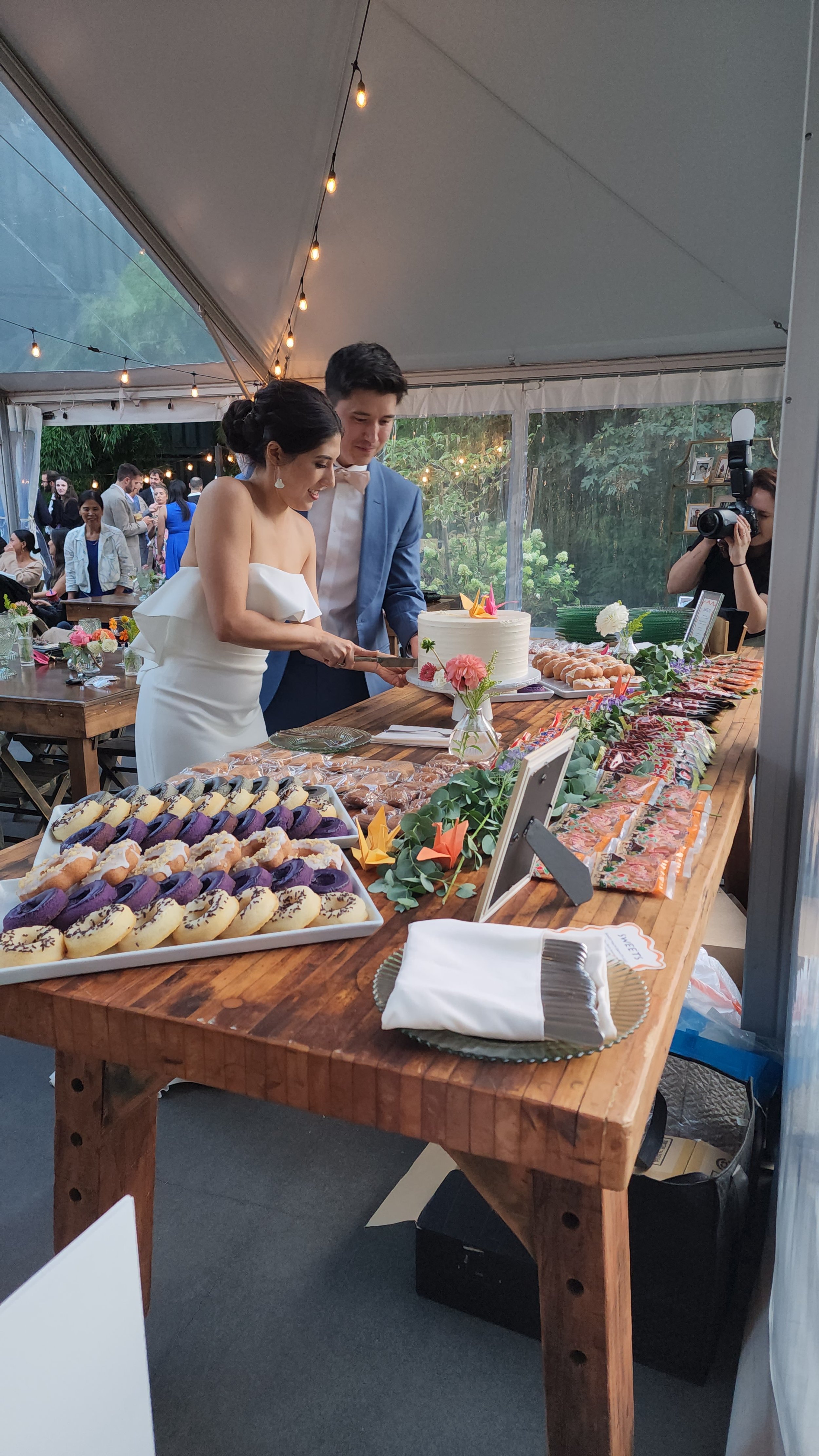 Couple cutting a wedding cake at a reception with a photographer capturing the moment. There's a dessert table with various pastries and decorations, under a tent with string lights.