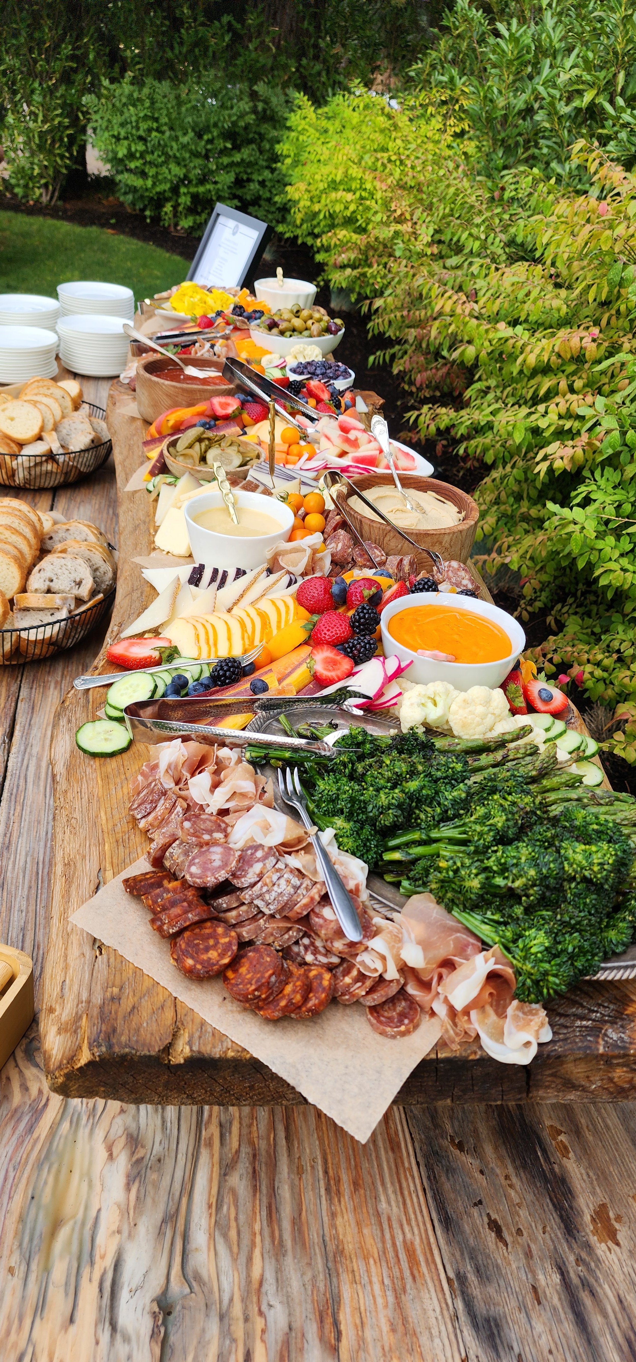 Charcuterie board with meats, cheese, fruits, and vegetables on a wooden table outdoors, surrounded by greenery.