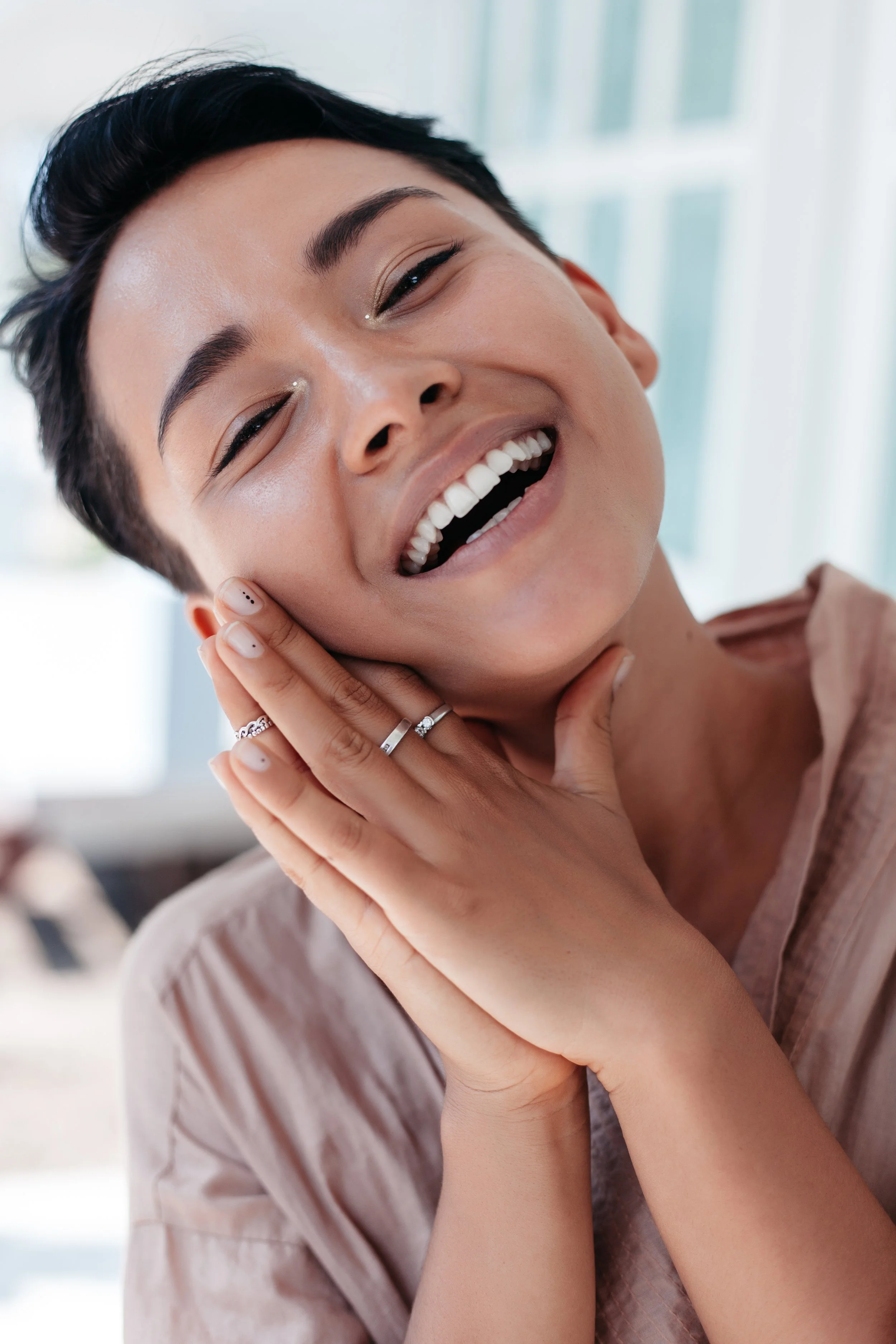 Une femme asiatique métisse au maquillage subtil sourit à l’appareil photo et pose sa tête sur ses mains.
