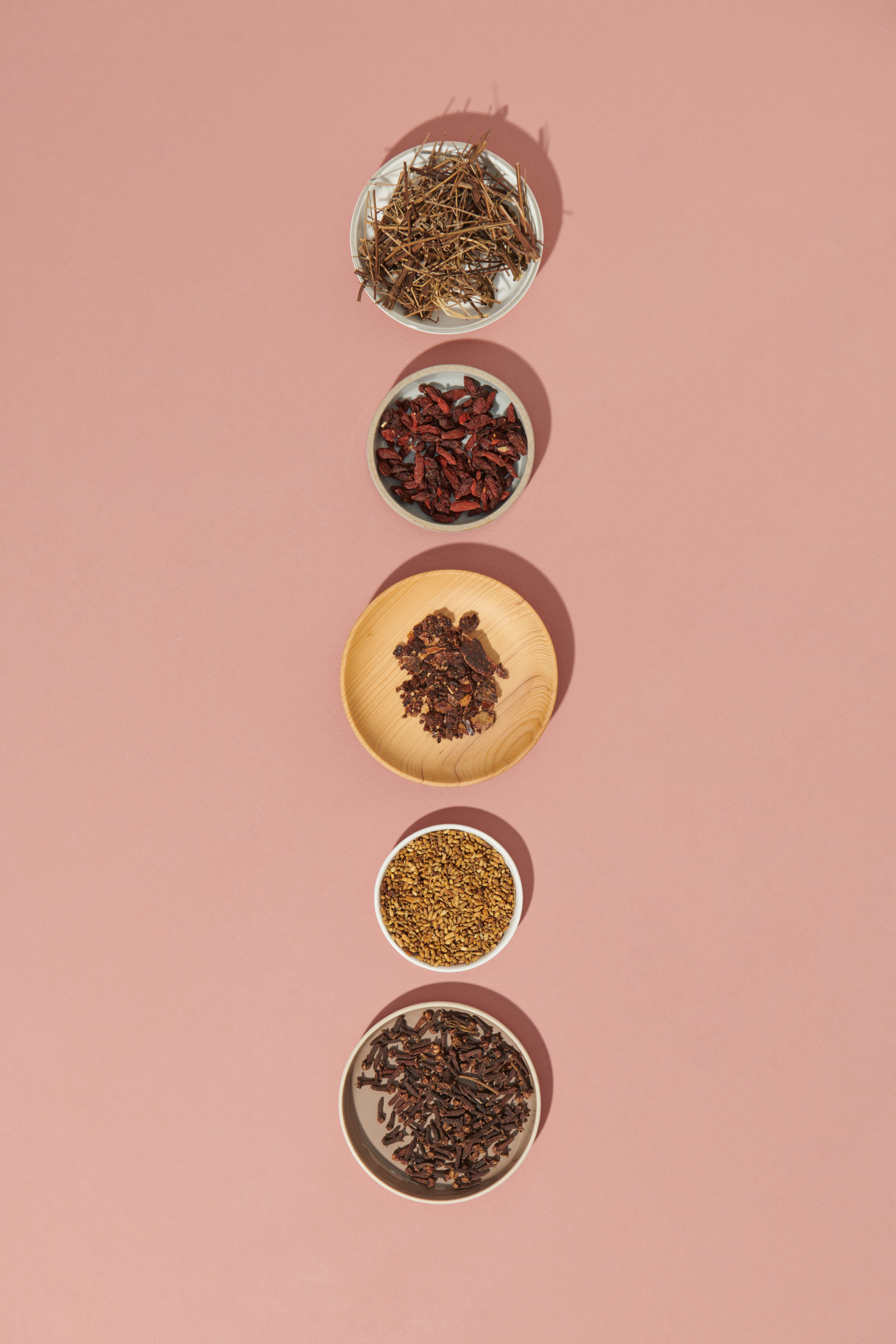 Une série d’ingrédients à base de plantes placés dans de petites assiettes en ligne verticale sur un fond rose.