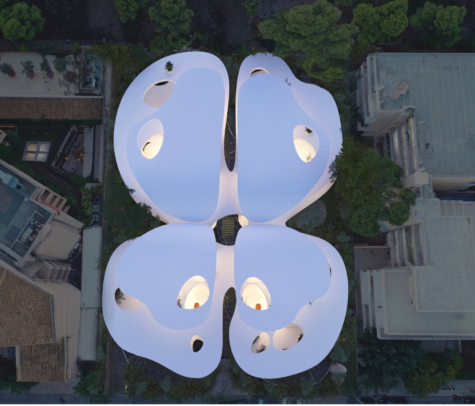 Aerial view of a butterfly-shaped modern architectural building with white walls and circular cutouts, surrounded by streets, trees, and neighboring buildings at dusk.