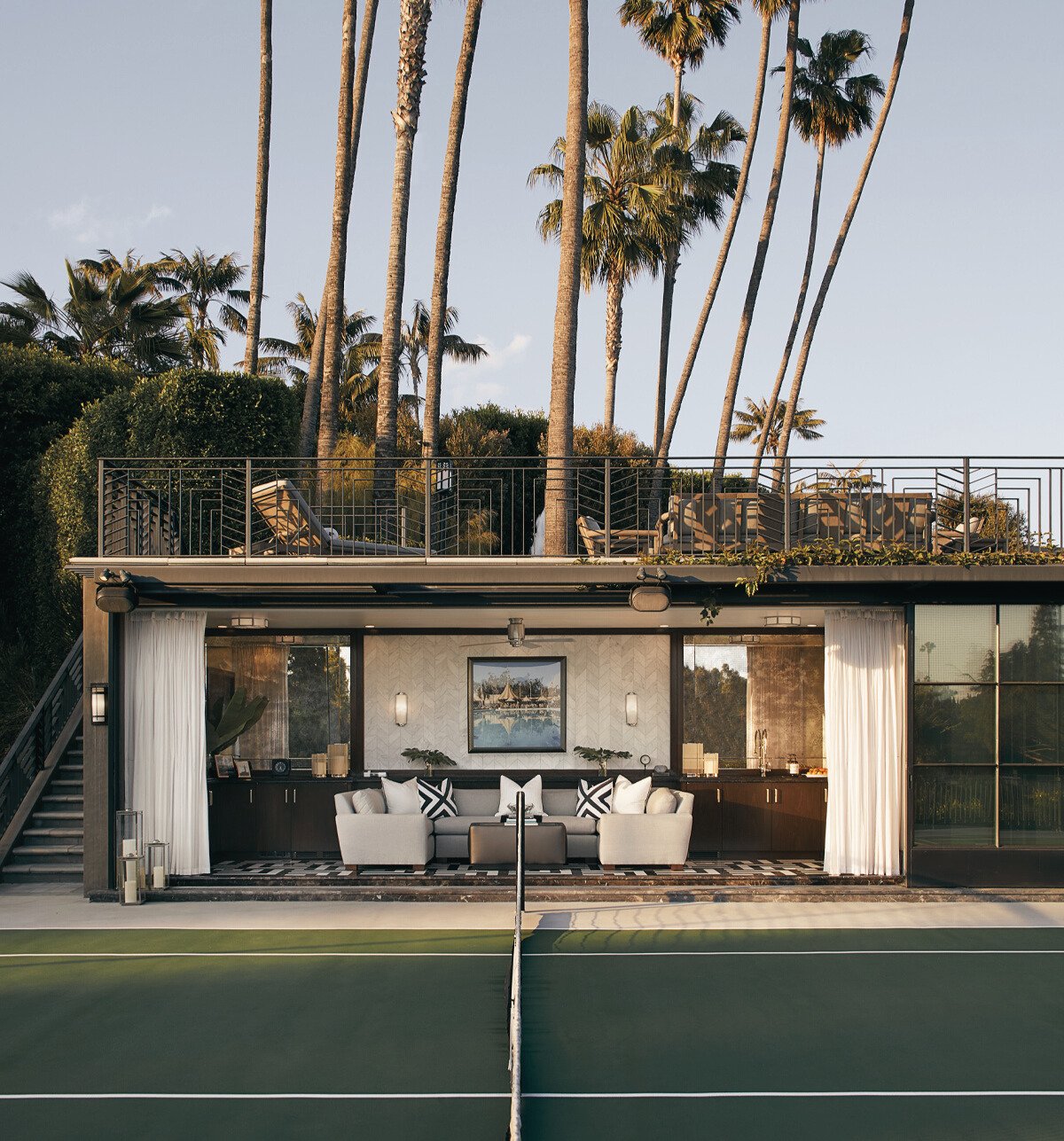A two-story house with an upper balcony surrounded by tall palm trees, and a ground-level outdoor living area with a sofa, pillows, and a painting on the wall, overlooking a tennis court.
