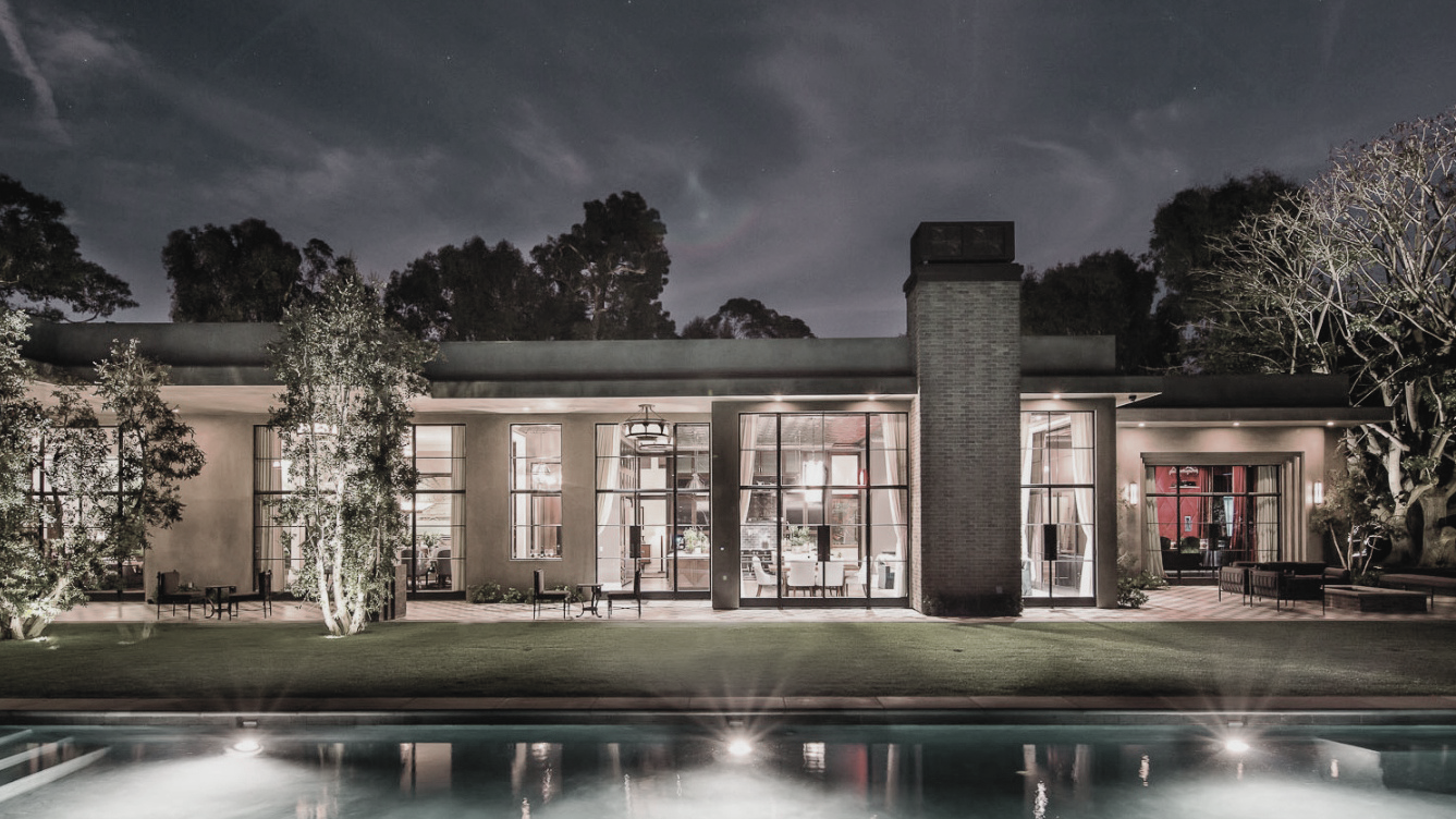 Nighttime view of a modern house with large glass windows, illuminated interiors, outdoor seating, and a swimming pool in the foreground.
