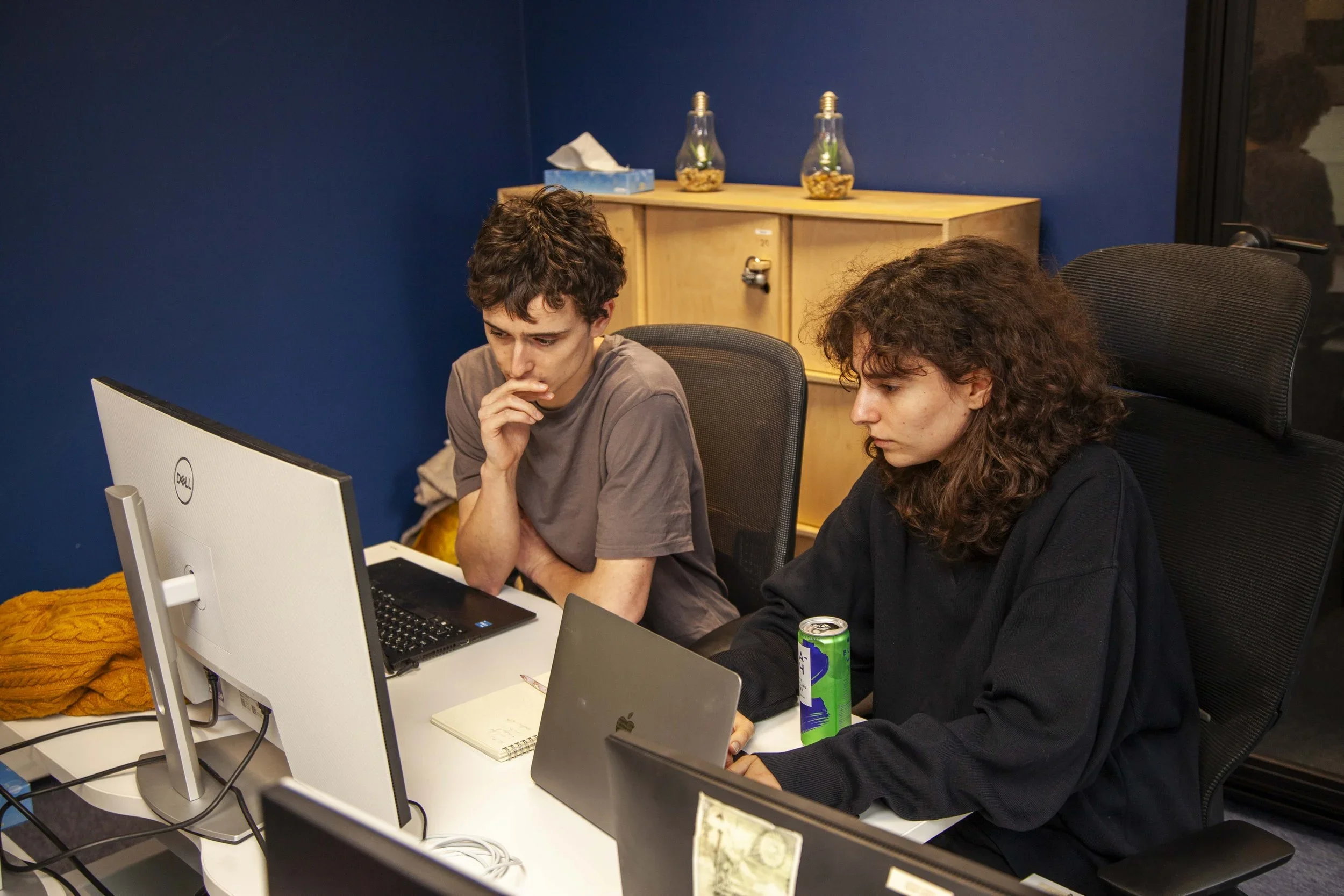 Two people working at a desk with laptops and a computer monitor in an office setting, with blue walls and a shelf in the background.