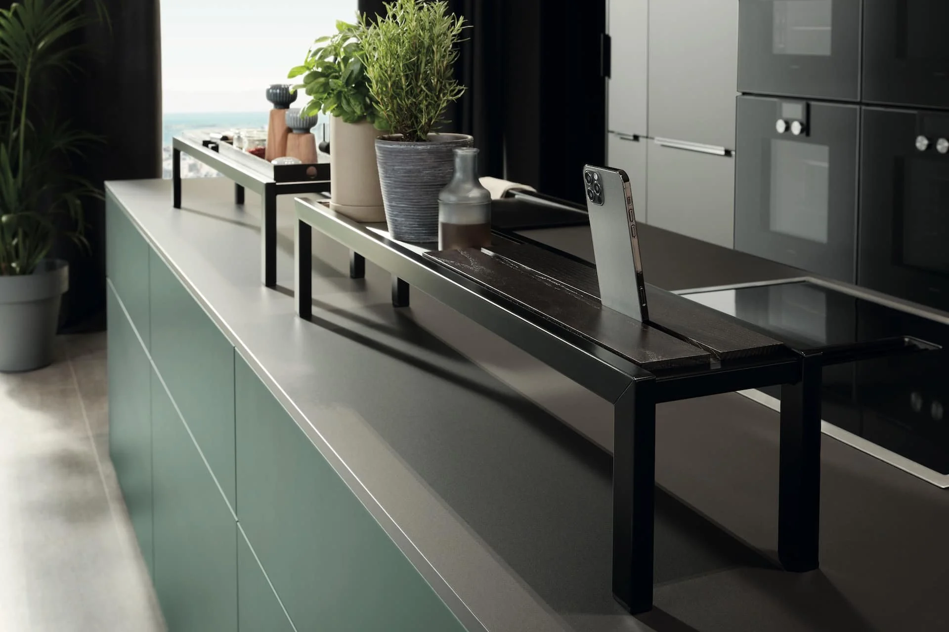 Modern kitchen countertop with plants, a smartphone, and a black tray with bottles and bowls.