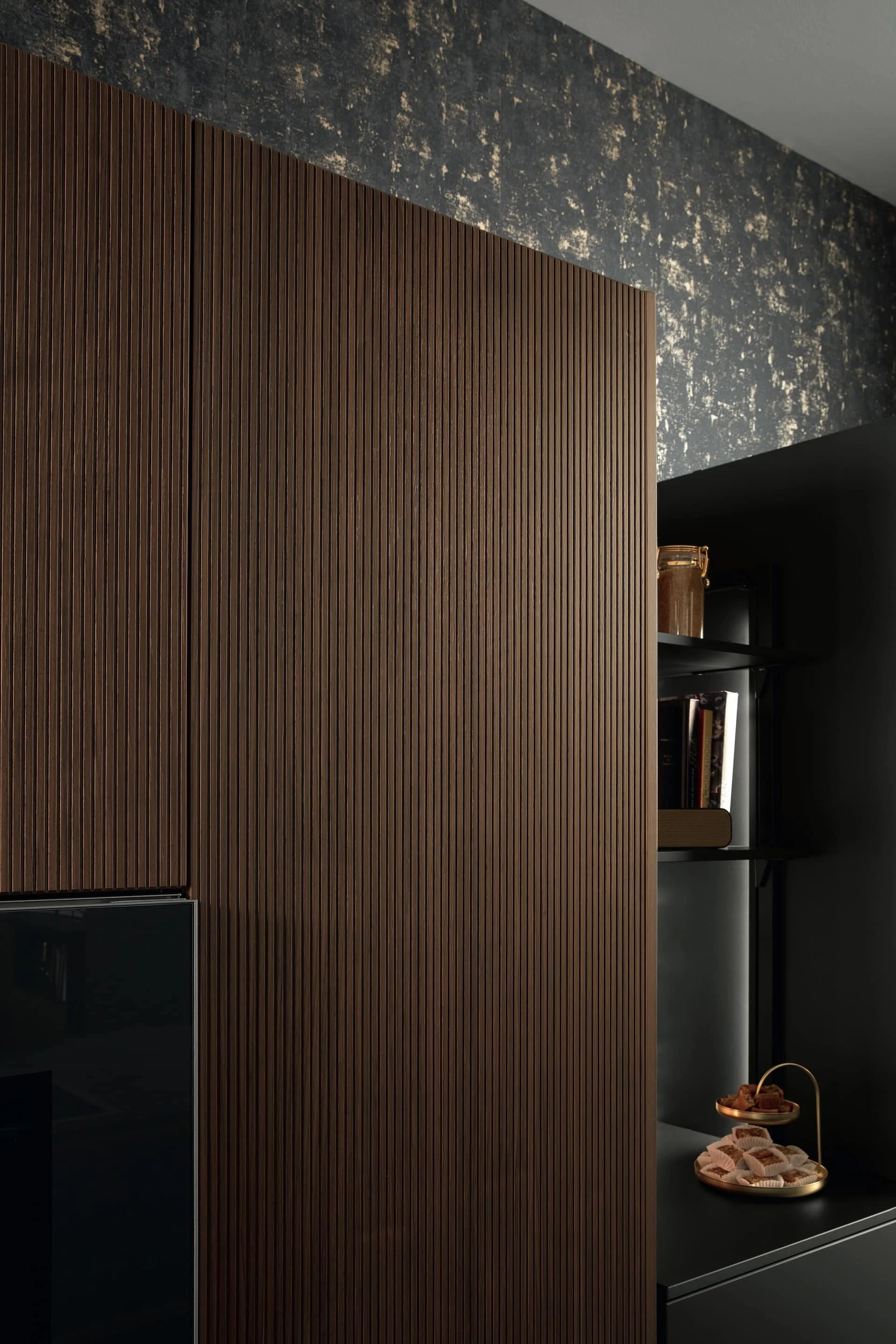 Interior scene with a large wooden cabinet with vertical grooves, dark shelving with books and decorative items, and a black surface with a tray of snacks, all in a modern style.