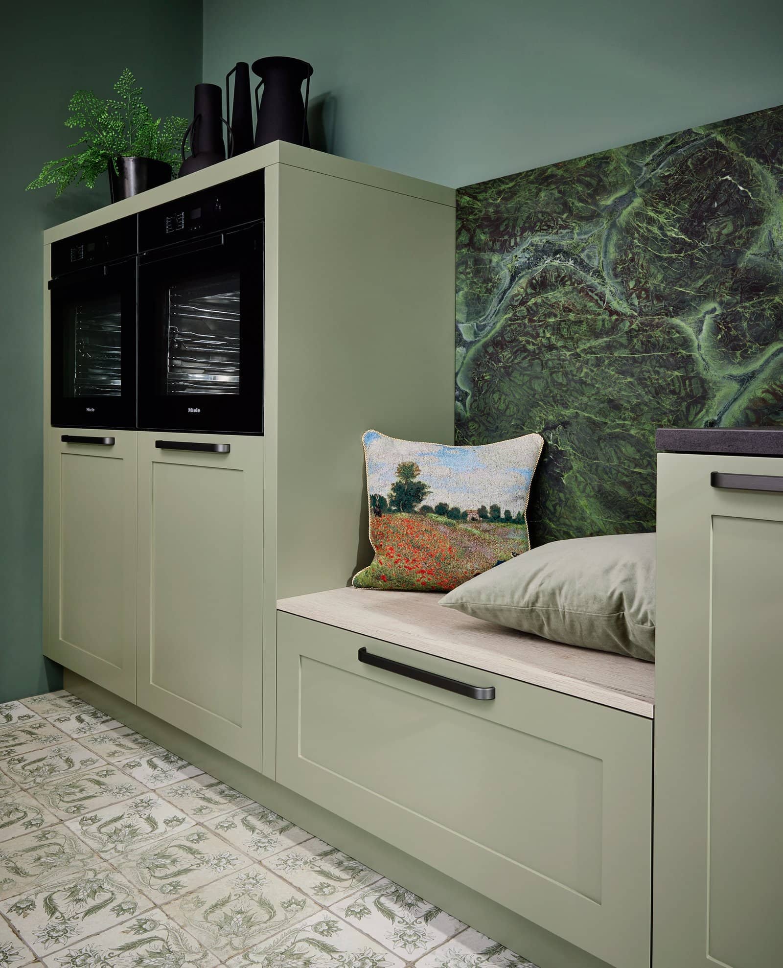 Corner of a kitchen with green cabinetry, built-in black ovens, a green accent wall with a nature mural, a cushioned bench with pillows, and decorative black vases and plants on top.