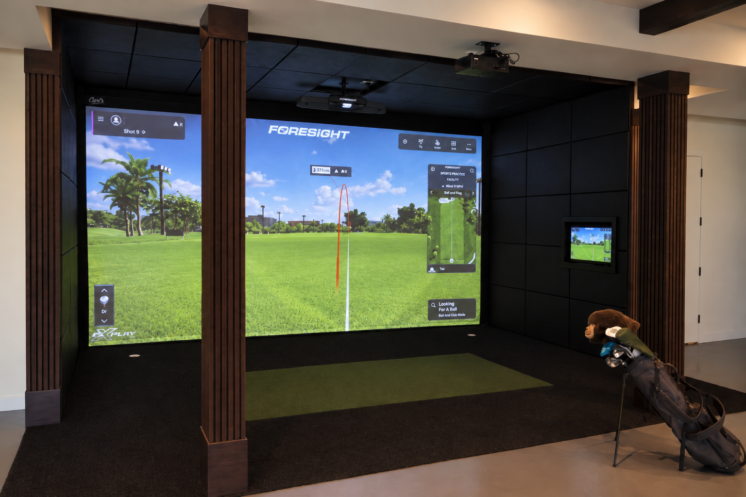 Indoor golf simulation featuring a large screen displaying a golf course, golf clubs and a golf bag on a stand in front, and a smaller screen on the wall.