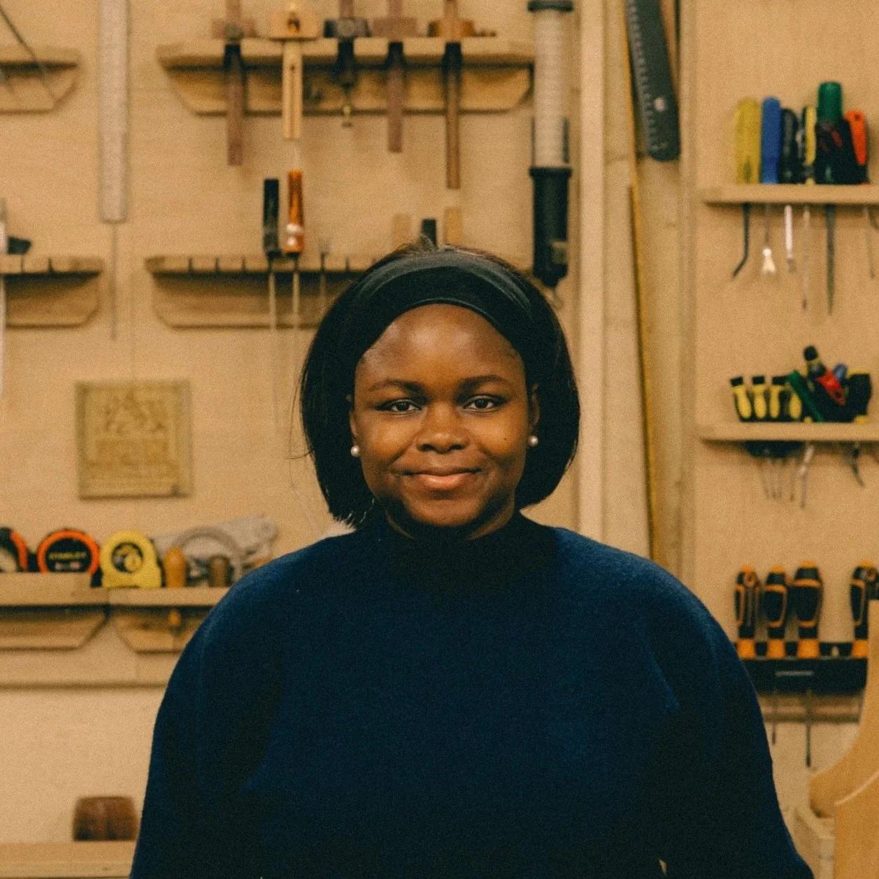 A woman with long wavy reddish-brown hair smiling in a woodworking shop with tools and wooden shelves in the background.