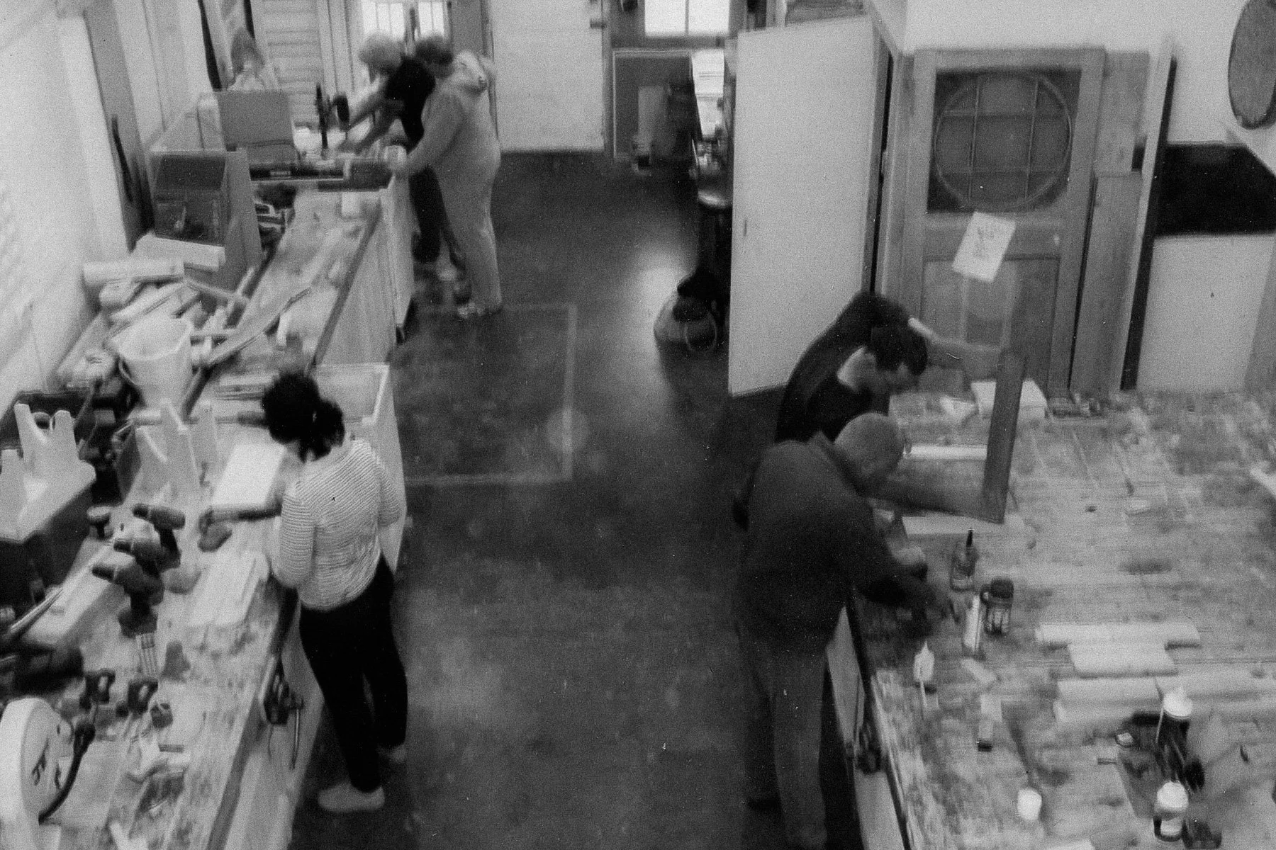 A black and white photo of a woodworking workshop with several people working on projects. Some are cutting or sanding wood, and the workspace is filled with tools, wood pieces, and equipment.