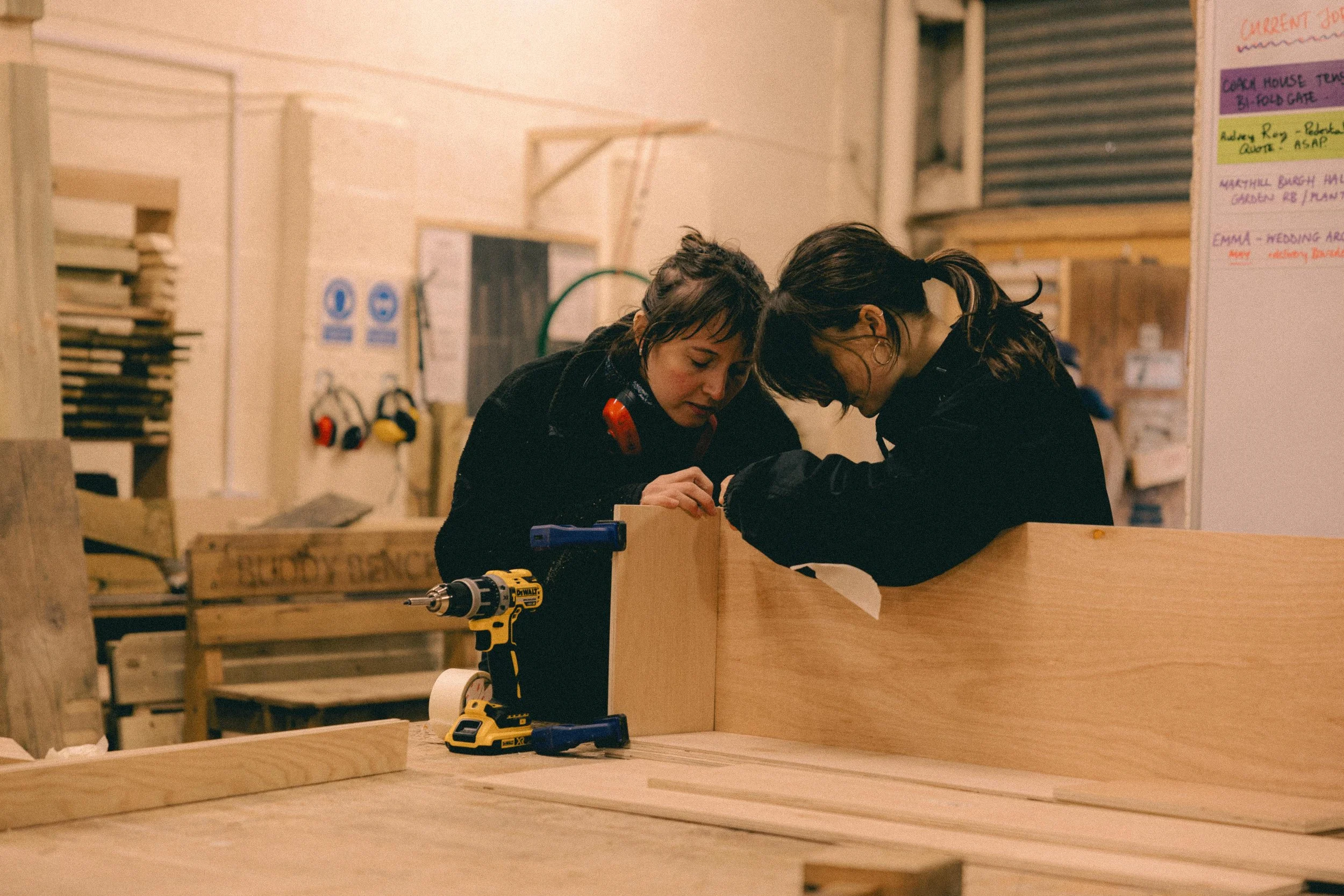 Two women working together on a woodworking project in a woodworking shop with tools and materials around them.