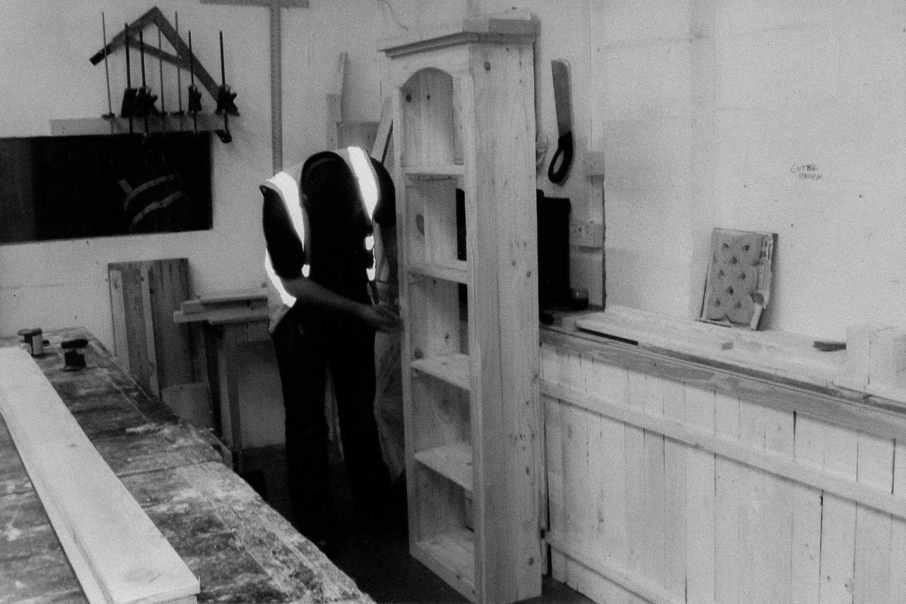 A person in a workshop surrounded by woodworking tools and furniture. The person is standing next to a wooden bookshelf, with shelves and a cabinet. The workspace includes various wood pieces, a workbench, and tools on the wall, such as saws and clam
