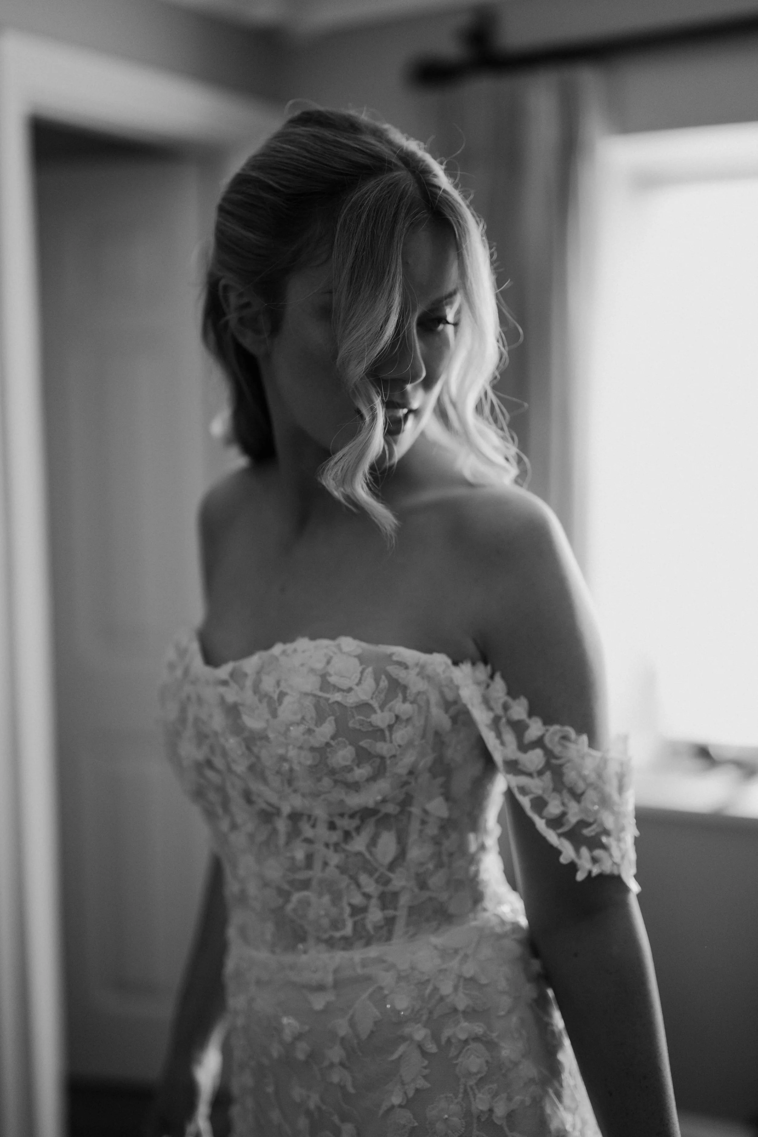 A black and white photo of a woman with wavy blonde hair, wearing a lace wedding dress, looking over her shoulder.