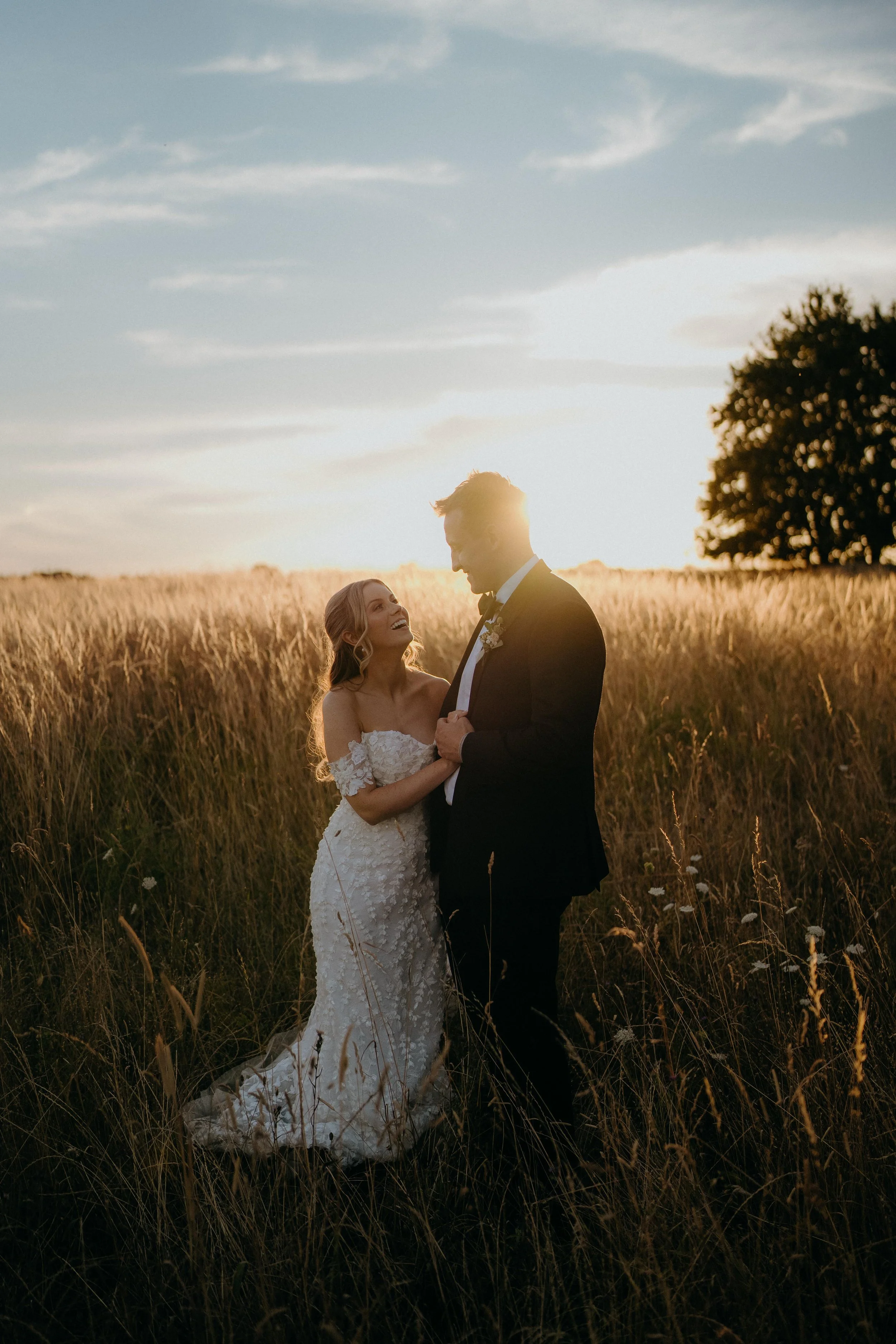 A bride and groom standing in a field of tall grass during sunset, smiling at each other, with the bride wearing a white lace wedding gown and the groom in a black tuxedo at Stone Barn in the cotswolds
