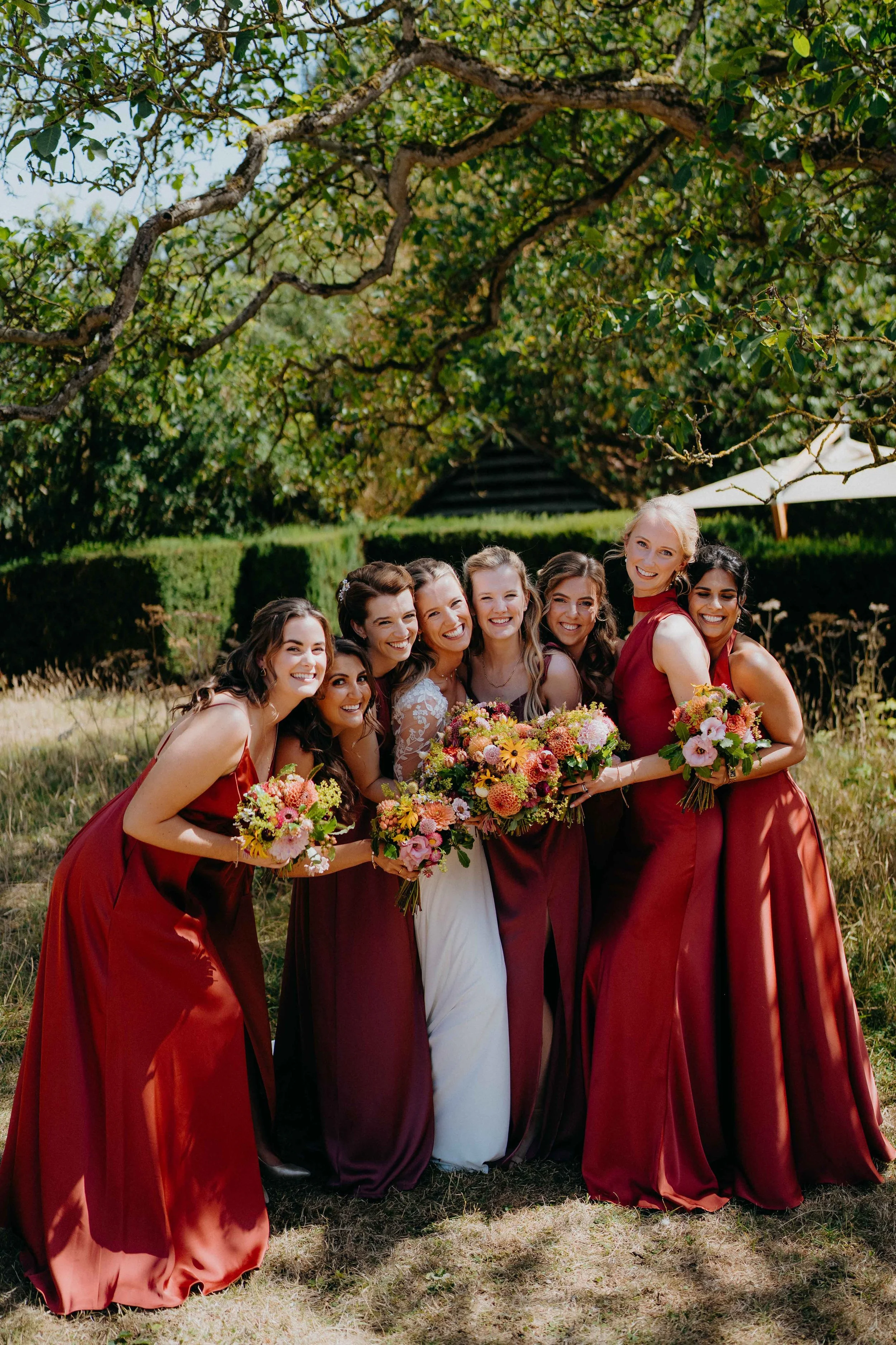 A group of women, including a bride in a white dress, standing outdoors under a tree during a wedding celebration. The women are holding colorful bouquets and wearing matching wine red dresses.