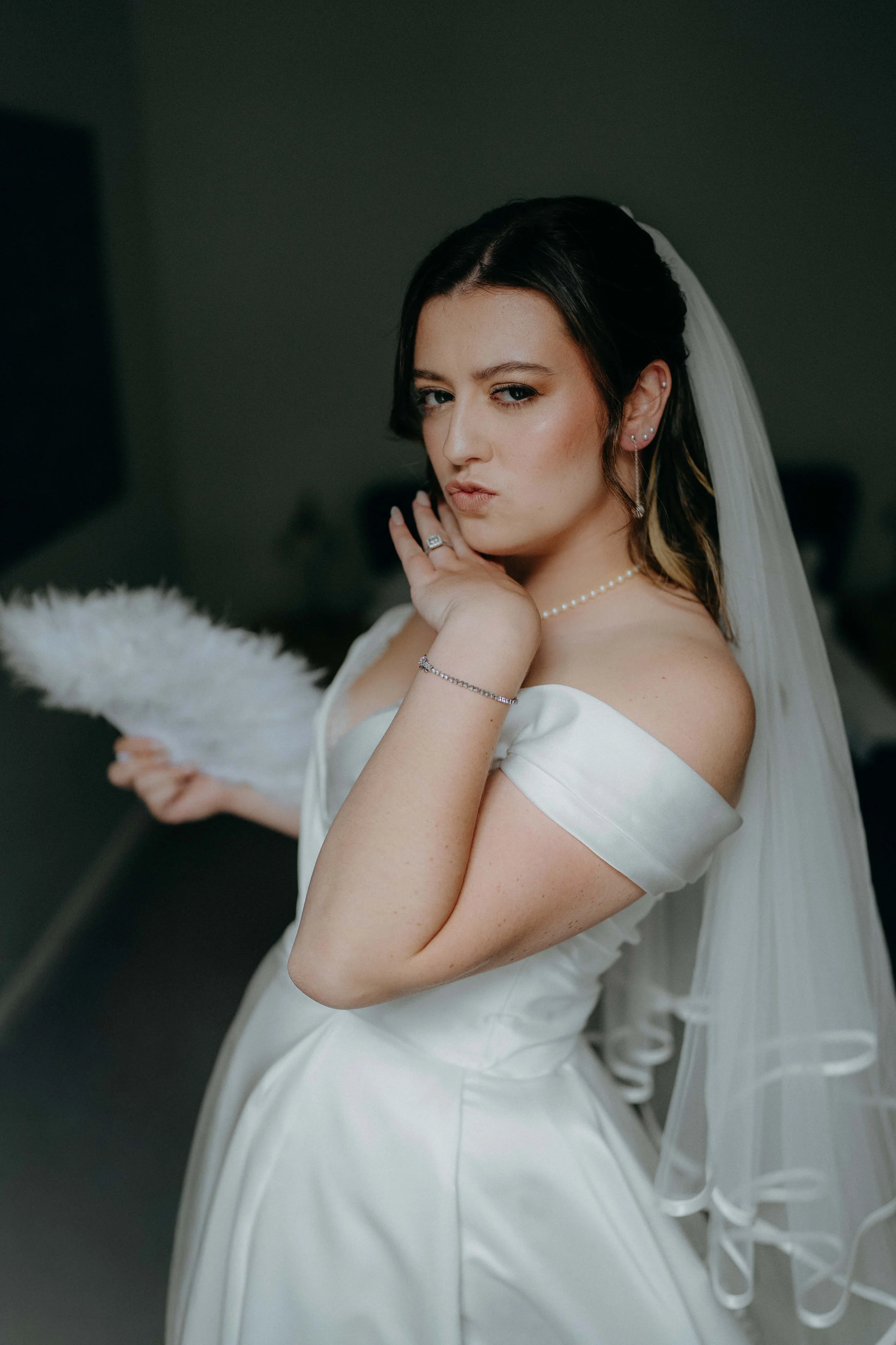 A woman dressed in a white wedding gown, with black hair and a veil, holding a feather duster and making a playful, sassy facial expression.